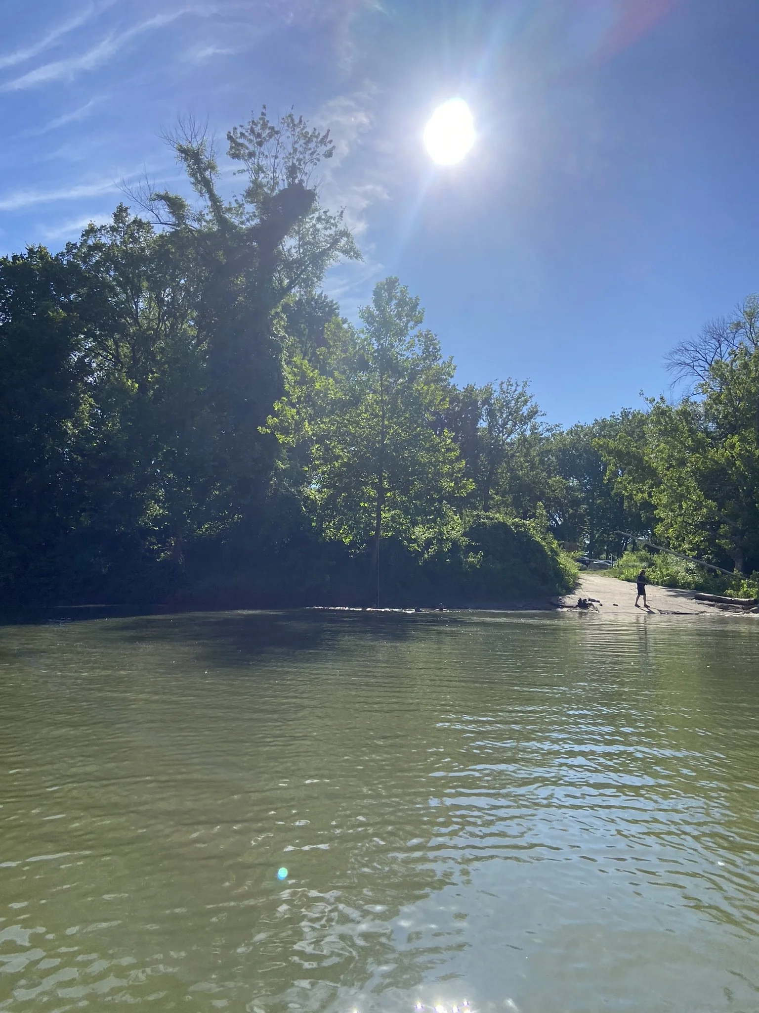 A peaceful outdoor scene of a river or lake with calm water, surrounded by green trees and bushes. The sun is shining brightly in a clear blue sky, creating a lens flare effect. There is a person in the distance on the shoreline with a fishing pole.