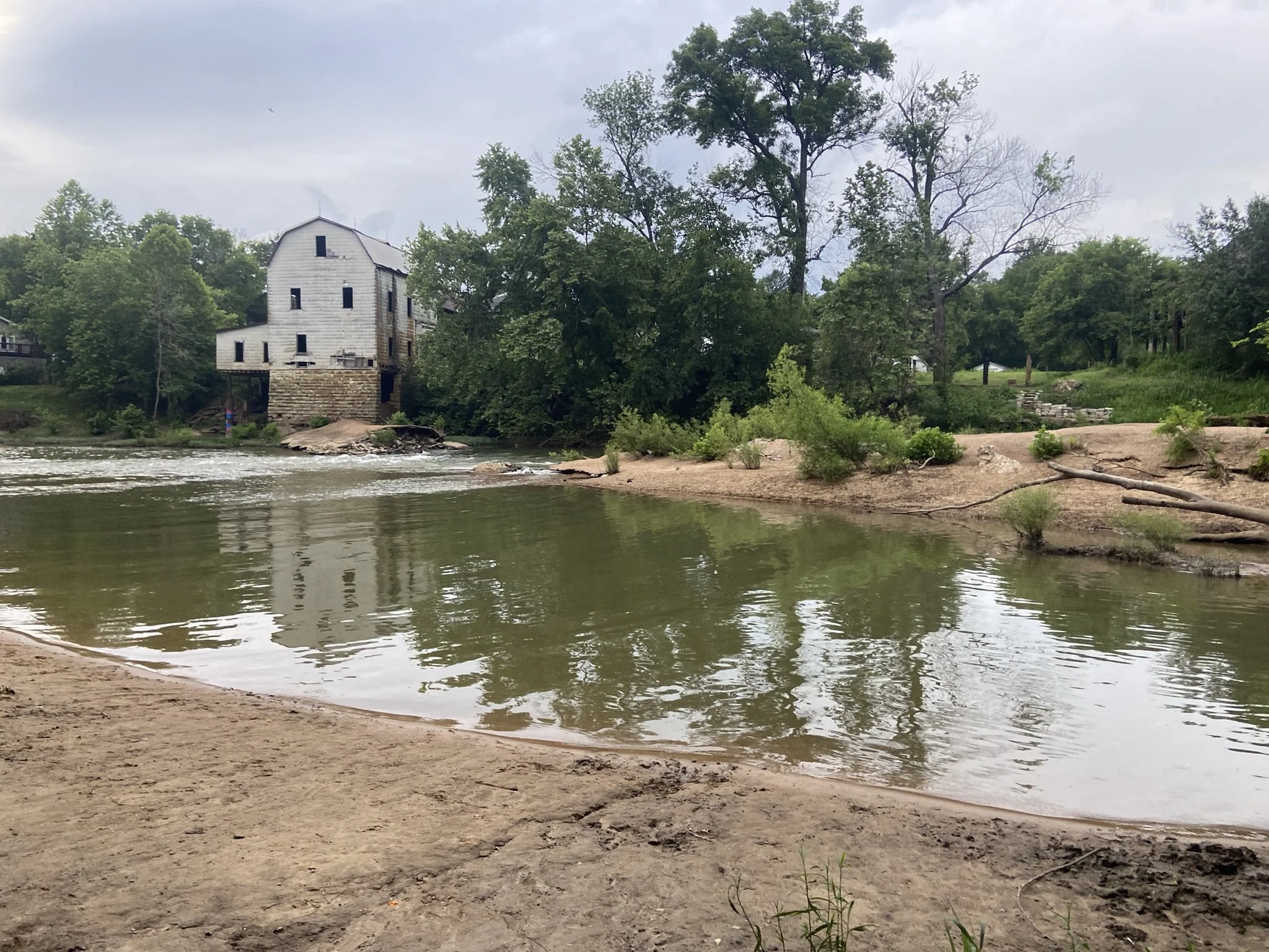 A river scene with a sandy shore in the foreground, trees along the riverbank, and an old weathered building on stilts with a stone foundation on the left side.