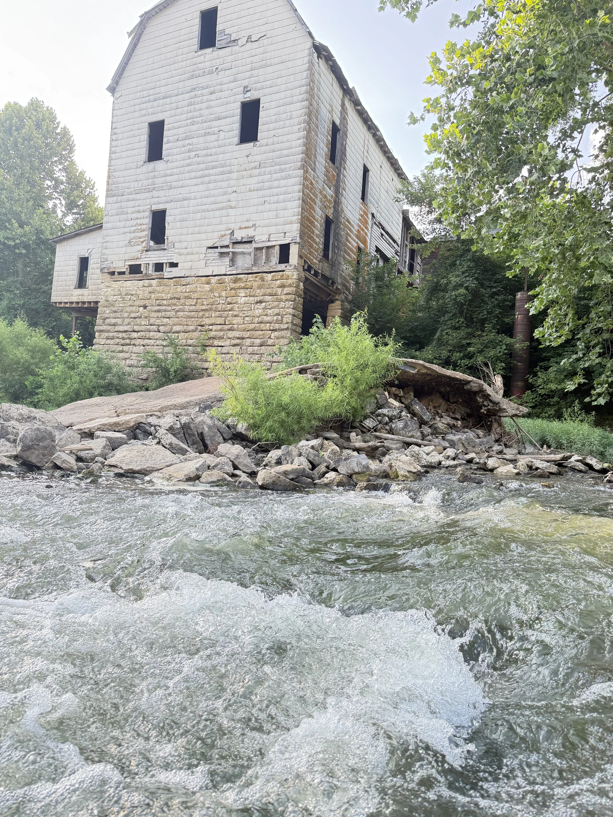 Old, abandoned house built on stone foundation, with damaged siding and missing windows, situated on rocky bank by flowing water surrounded by green trees.