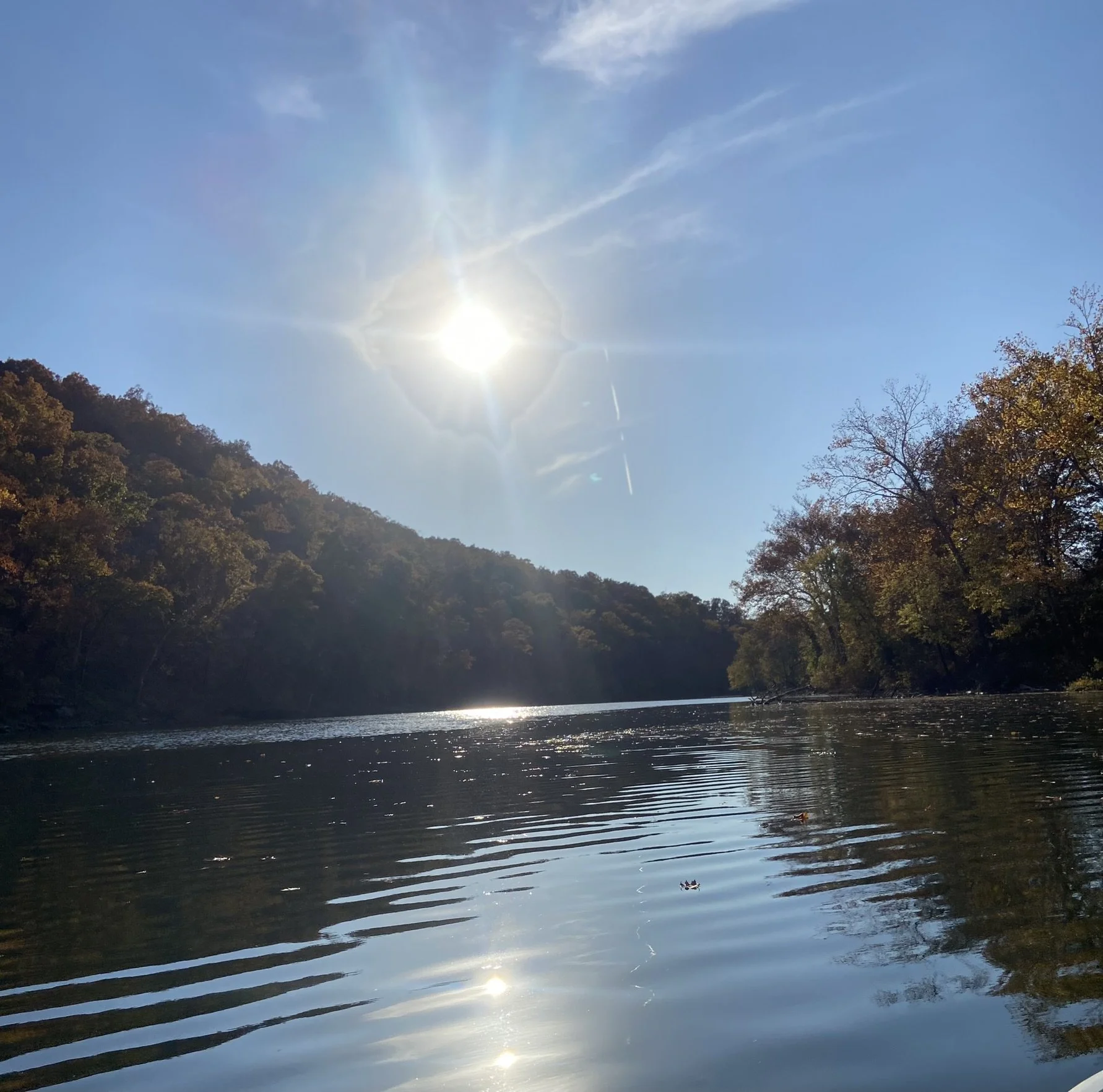 A bright sun over a river surrounded by trees with fall foliage, creating reflections on the water.