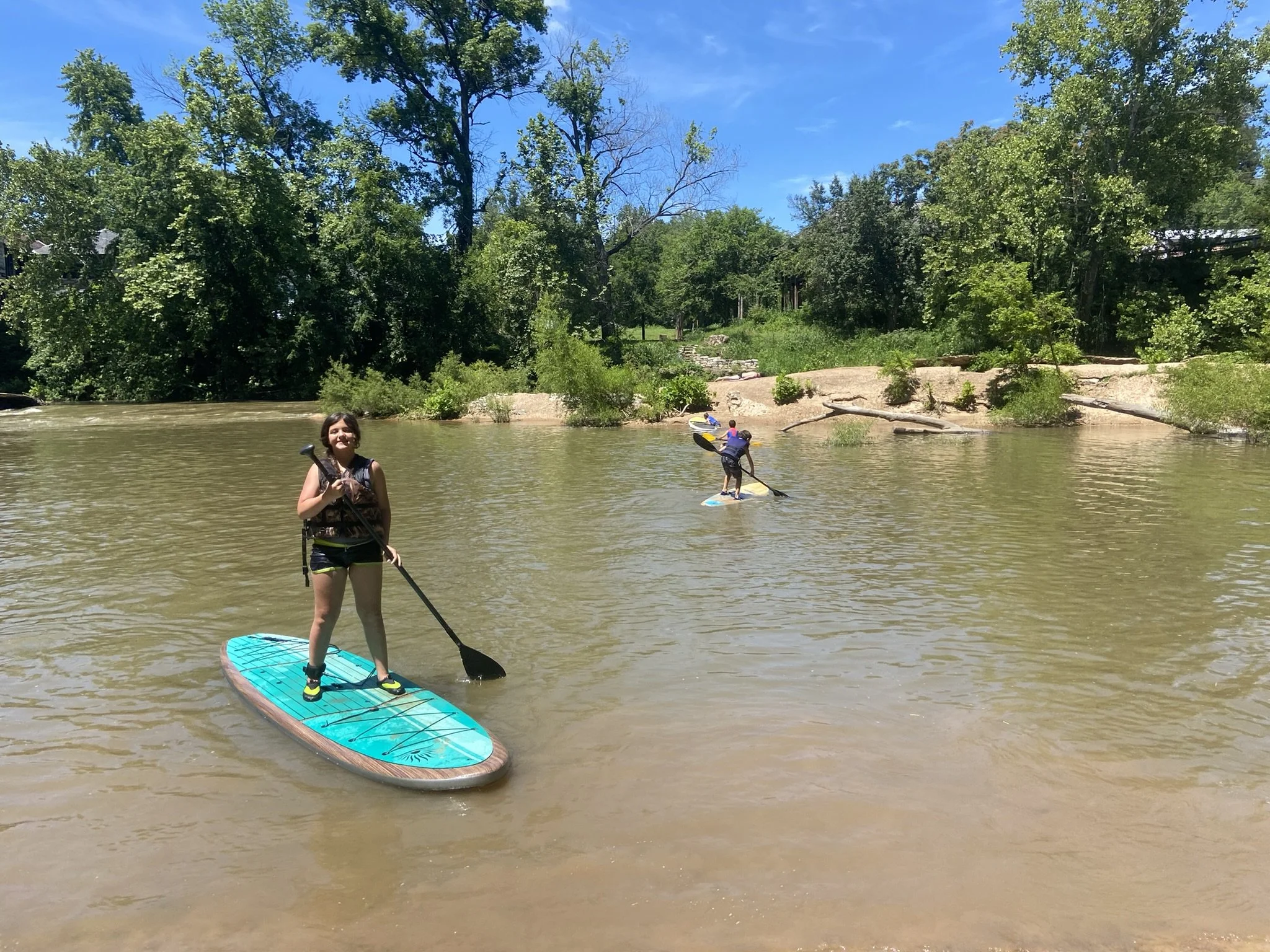 Children paddleboarding and kayaking on a river surrounded by green trees and a sandy shoreline on a sunny day.