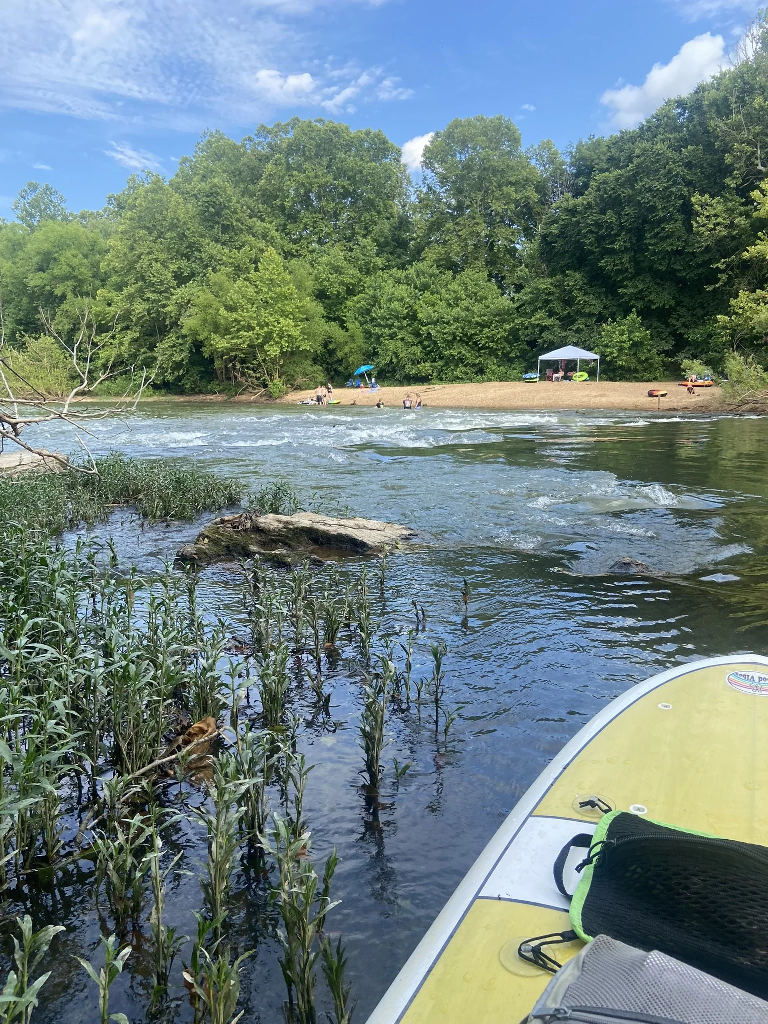 A calm river surrounded by lush green trees with a sandy beach on the farther side. On the beach, there is a white canopy tent, a blue umbrella, and a few people relaxing. The foreground shows part of a paddleboard floating on the river near some aquatic plants, with a black bag on the paddleboard. The sky is blue with some scattered clouds.