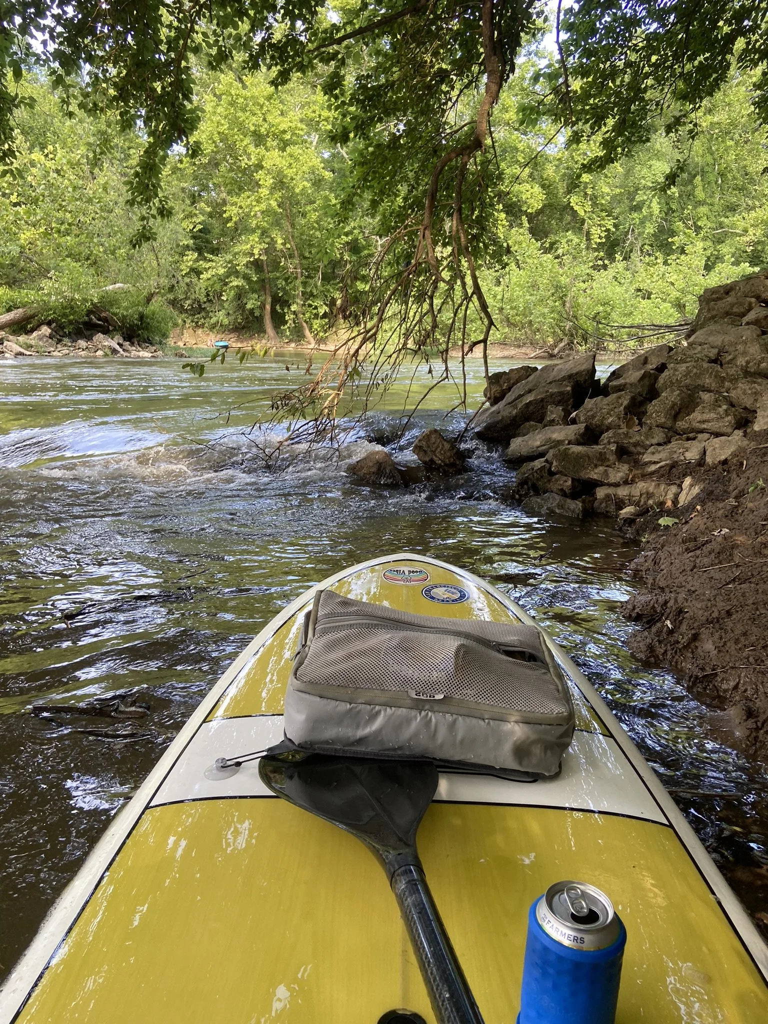 View from a yellow paddleboard on a river with trees and rocks along the shoreline, a paddle, a small bag, and a can of drink on the board.