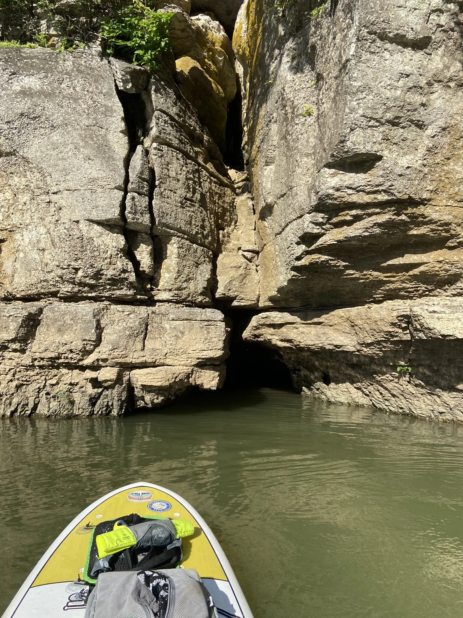 View from a paddleboard on a river, approaching a narrow canyon with large rock formations and a small dark opening in the rocks.