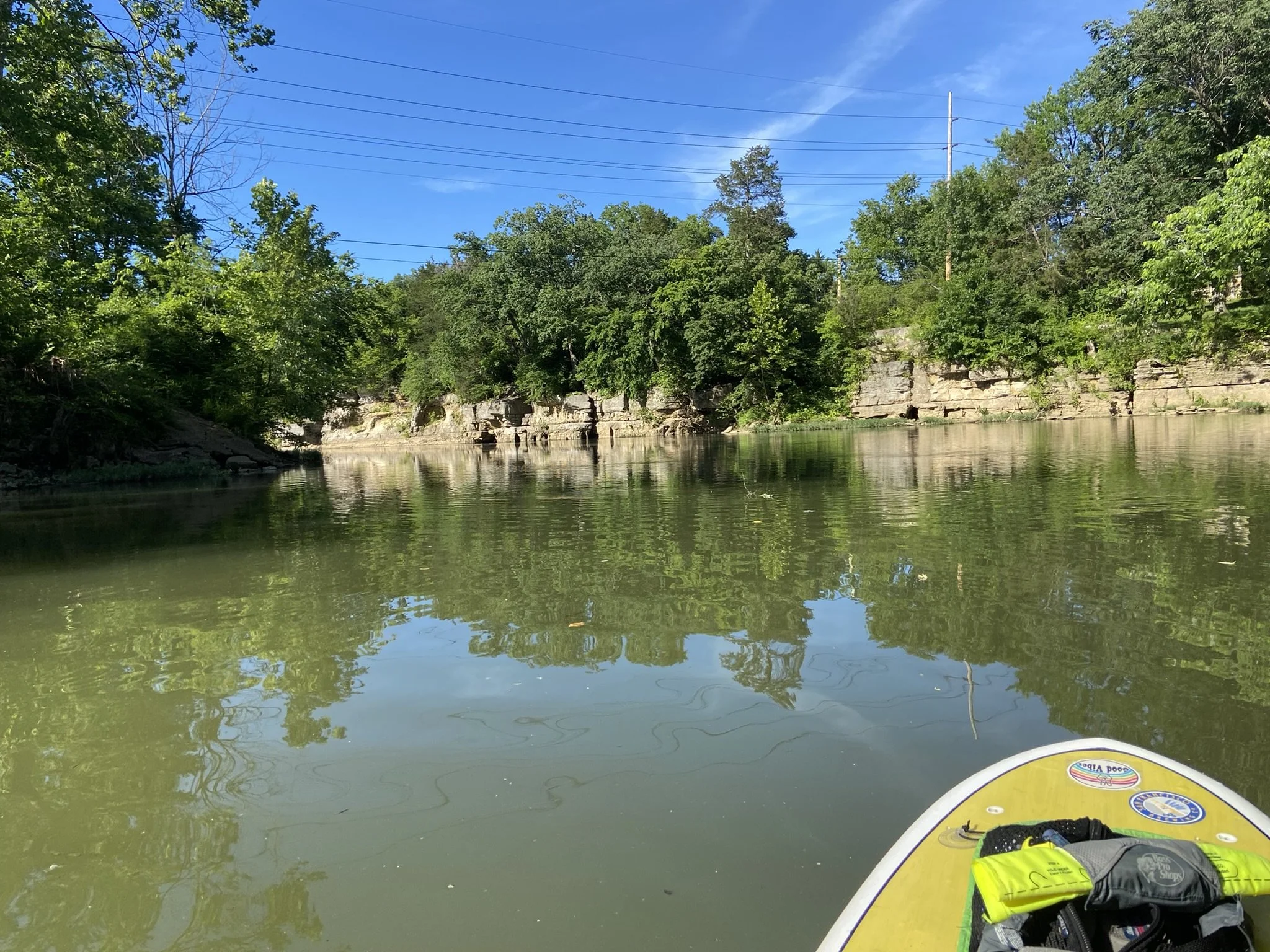 View from a paddleboard on a calm river with green trees and rocky cliffs along the shoreline under a blue sky.
