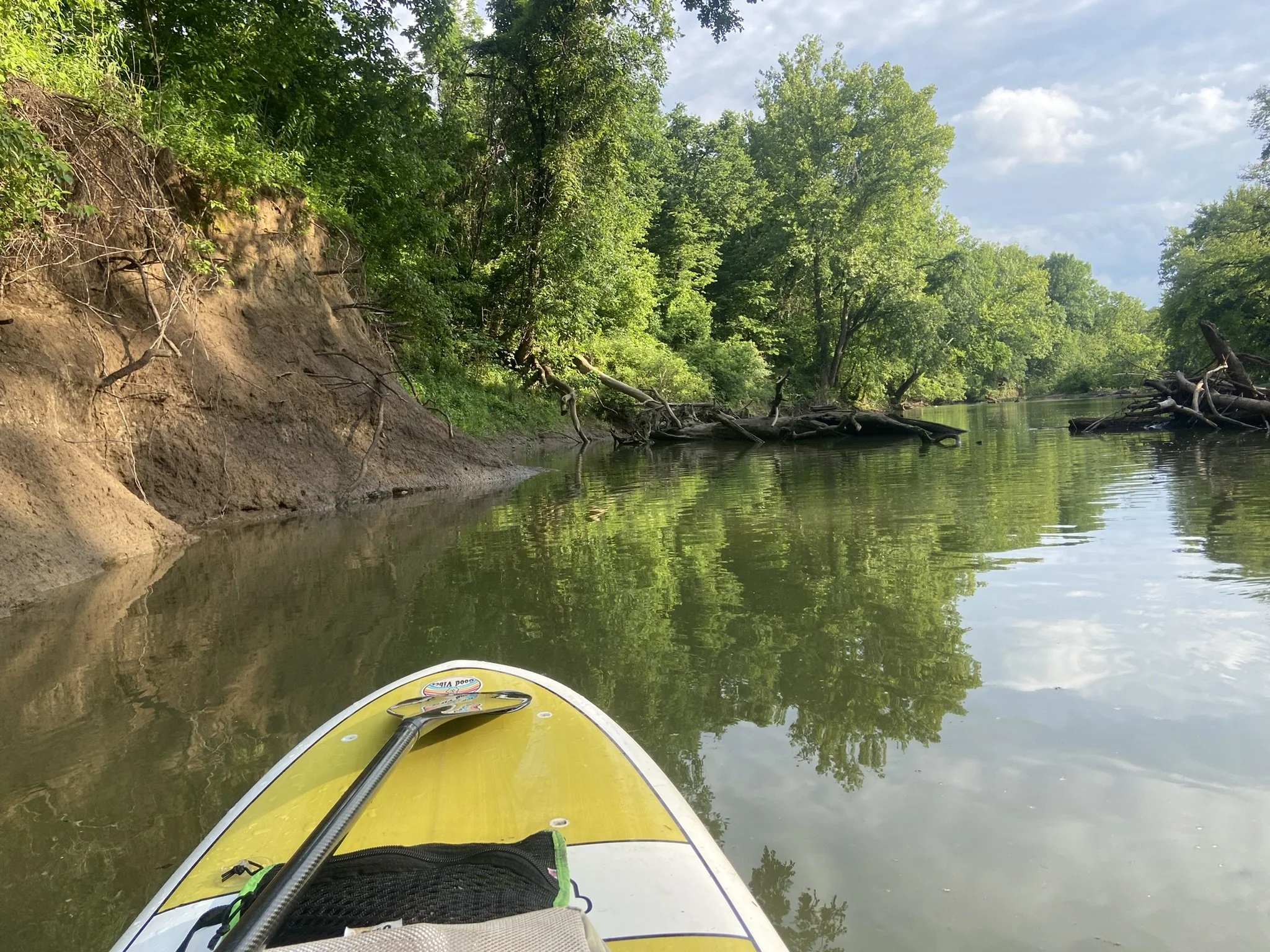 The image shows a calm river surrounded by lush green trees with a muddy bank on the left side. A yellow paddleboard with a paddle on top is in the foreground, floating on the water.