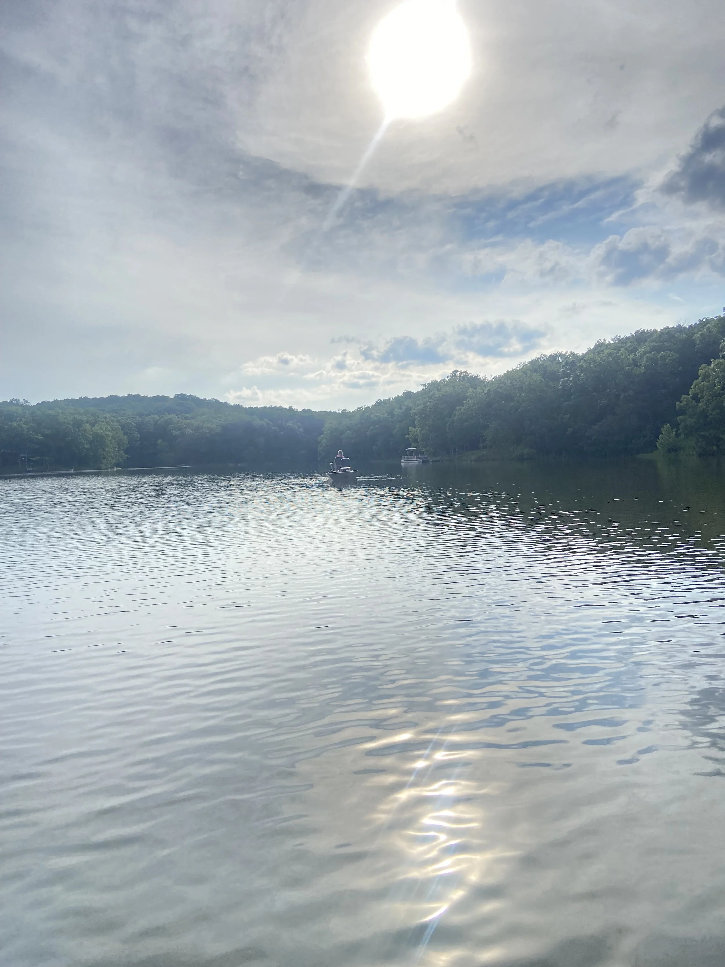 A calm river with two boats in the distance, surrounded by lush green trees under a partly cloudy sky, with the sun shining brightly overhead.