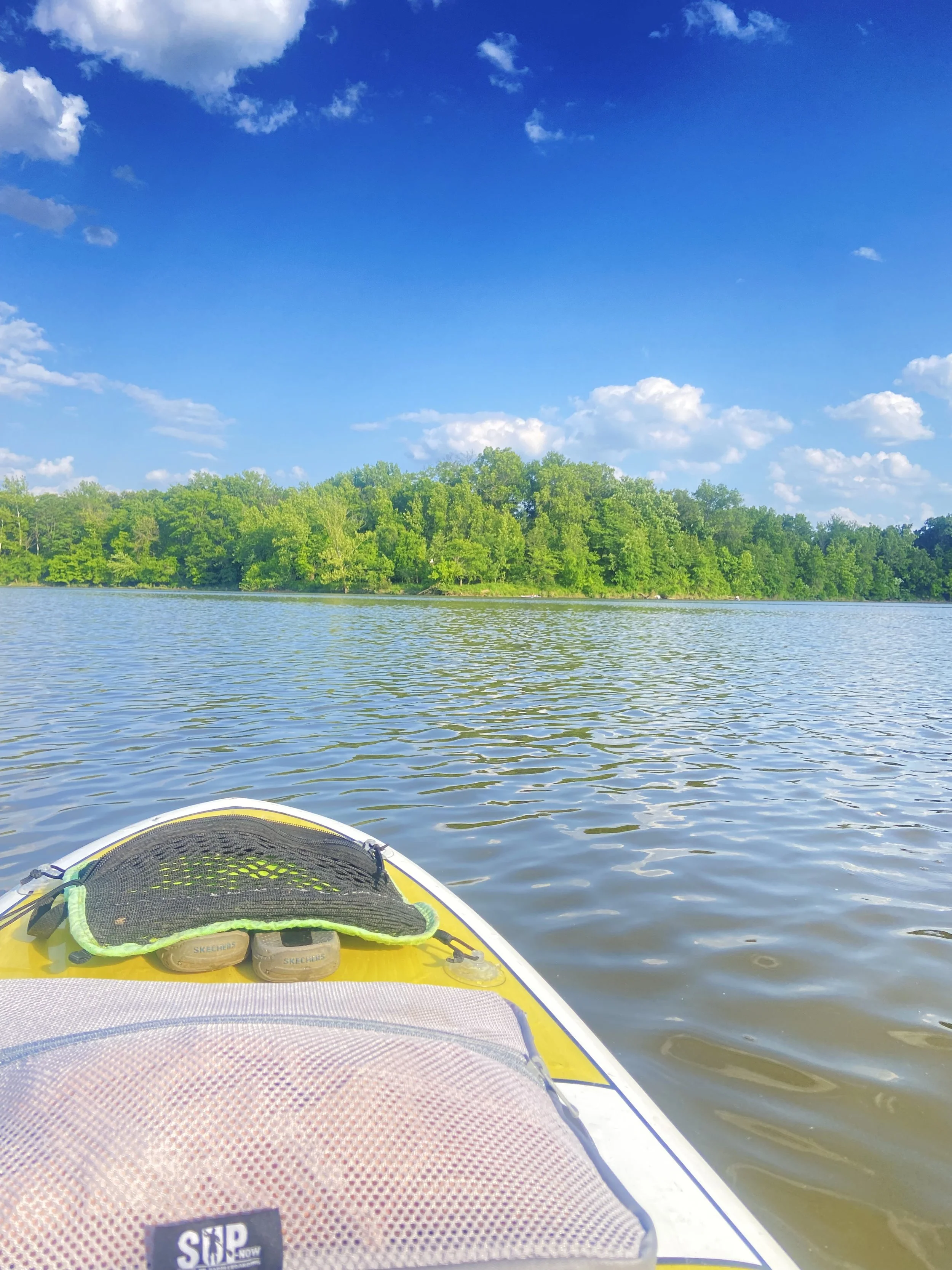 View from a SUP on a lake with a forested shoreline in the distance, a blue sky with scattered white clouds, and a yellow and white stand-up paddleboard in the foreground.