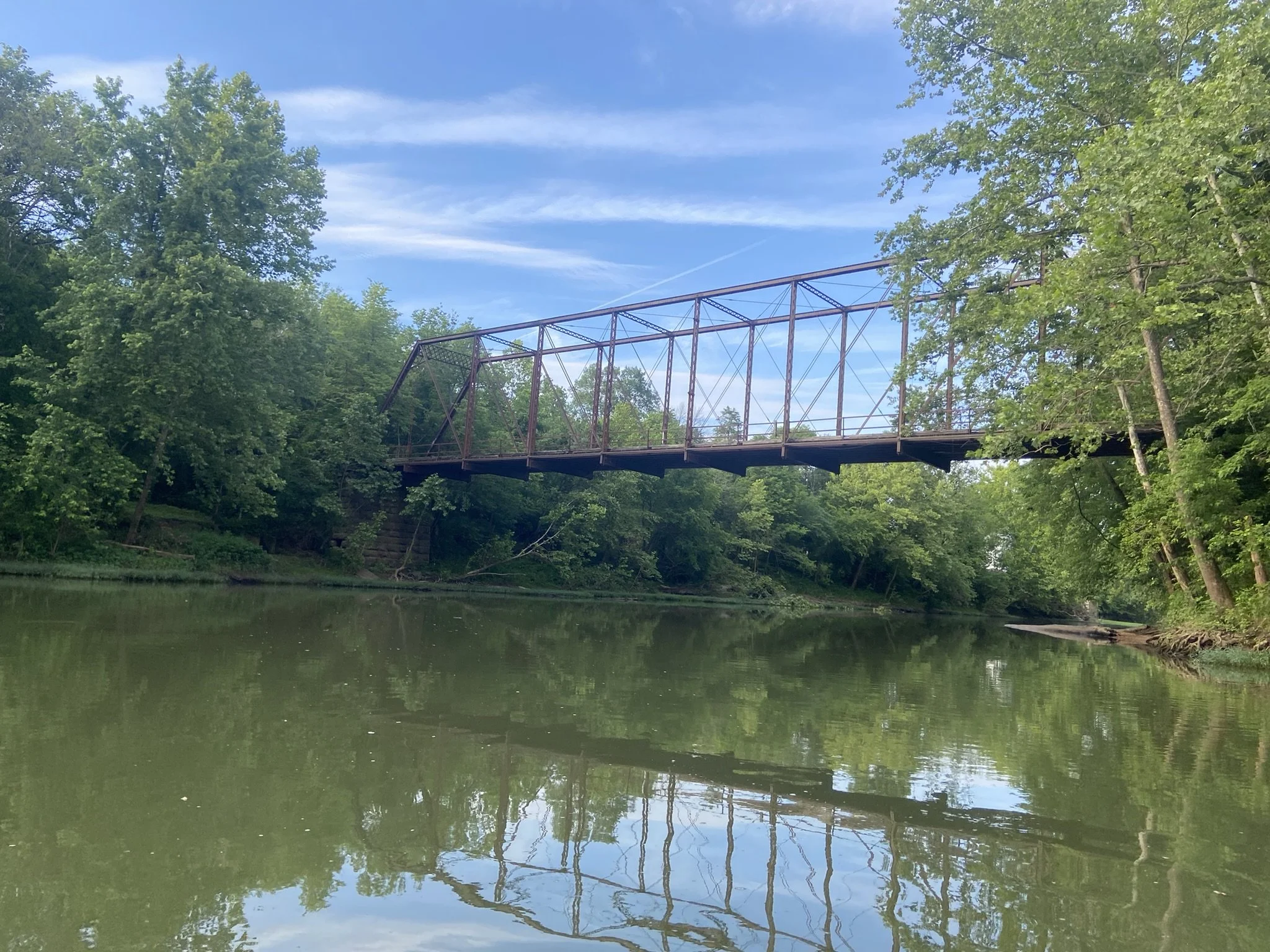 A rusty metal bridge over a calm river surrounded by lush green trees on a sunny day.