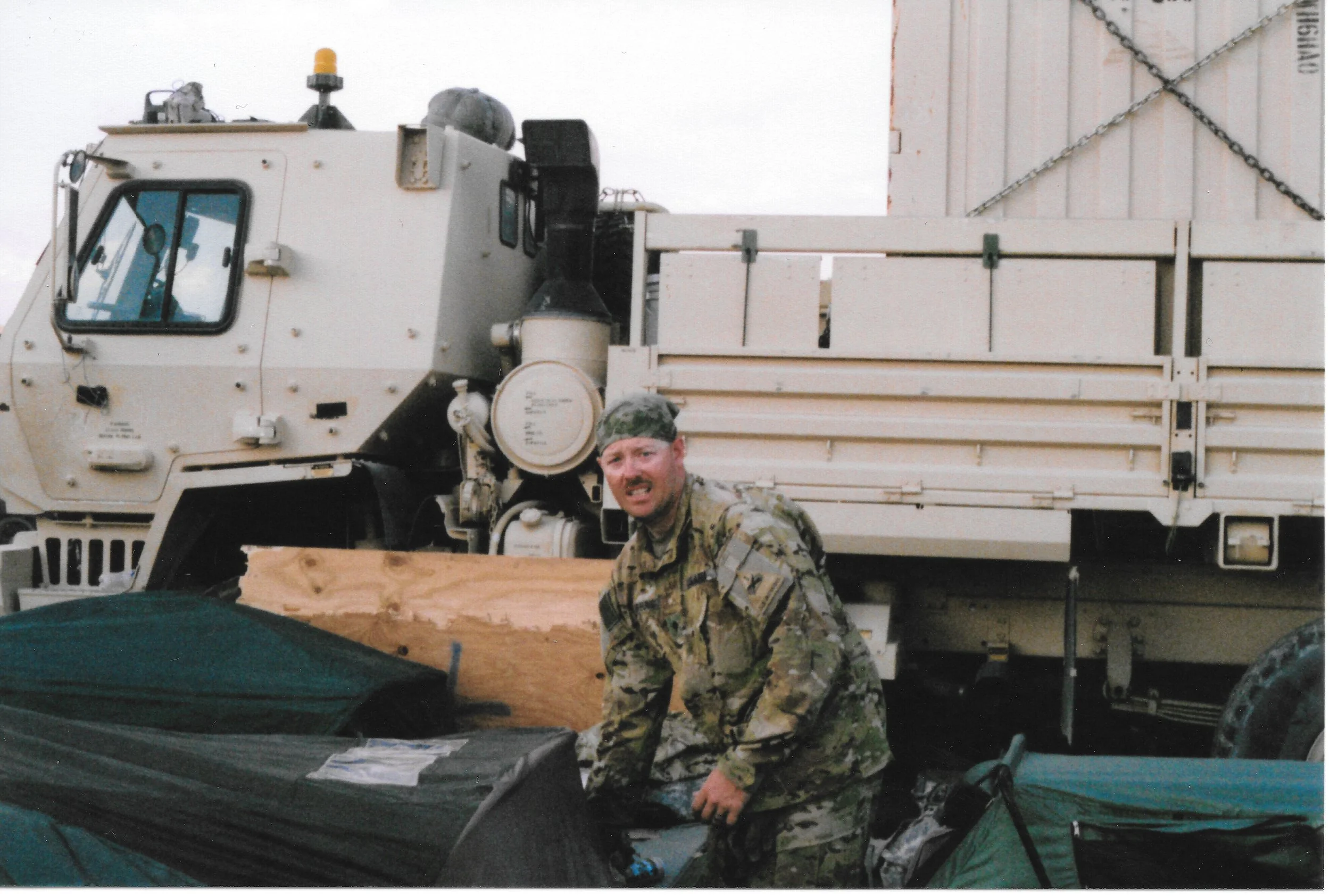A person in military camouflage uniform is smiling at the camera, standing in front of a large, white military vehicle with a flatbed and radar equipment on top. The person is also wearing a camouflage cap, and there is a wooden plank on the ground i