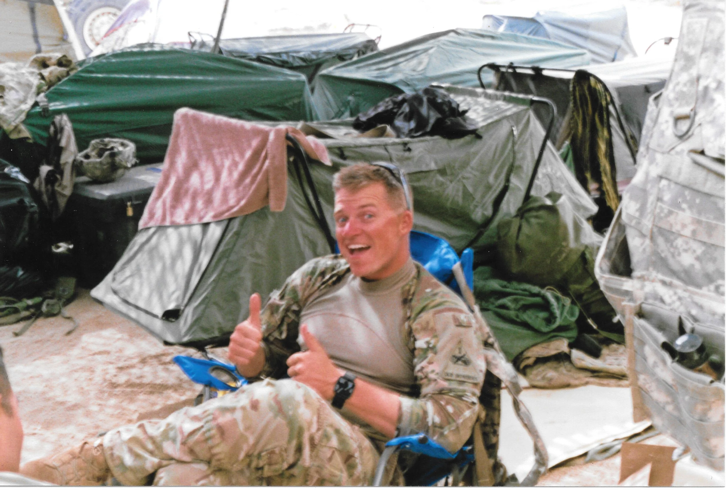 A man in military camouflage uniform smiling and giving a thumbs-up, sitting on a camping chair with a fully assembled tent and camping gear in the background.