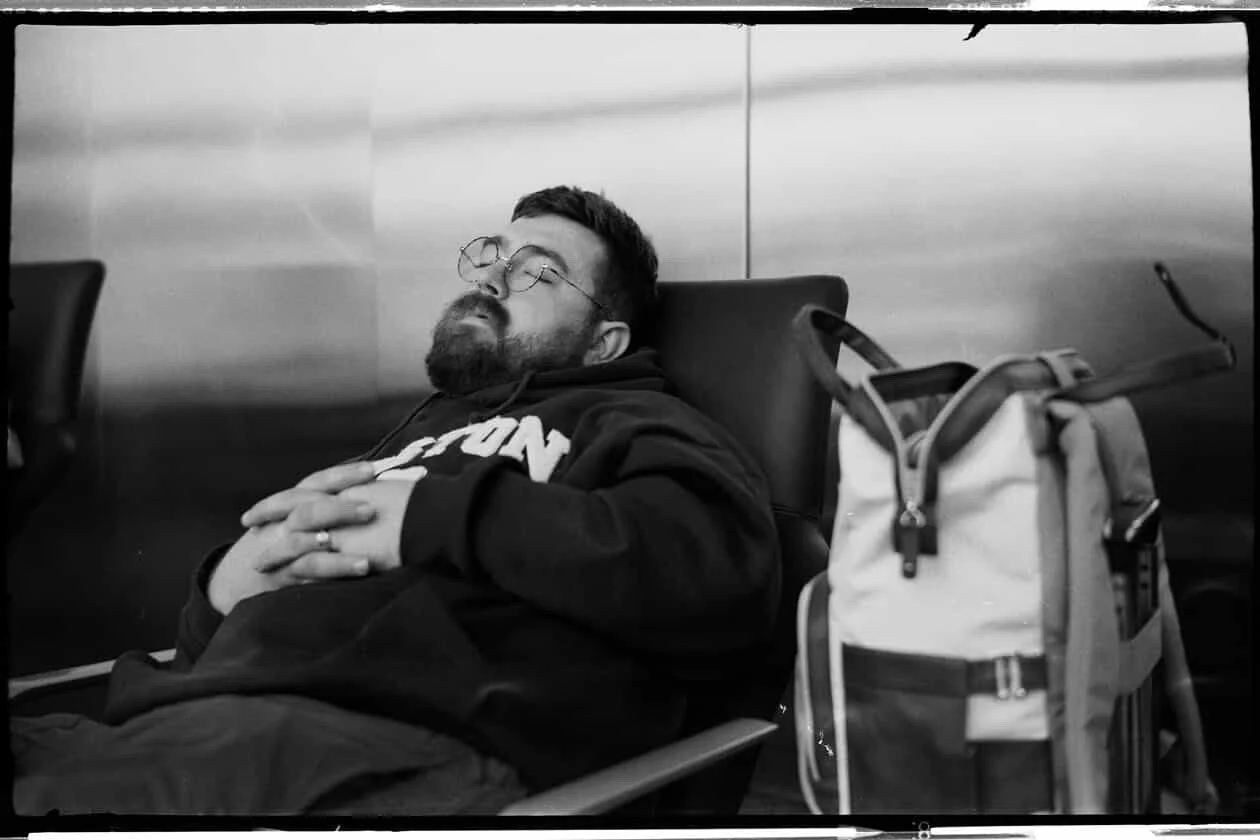 Man sleeping on airport chair with backpack beside him.