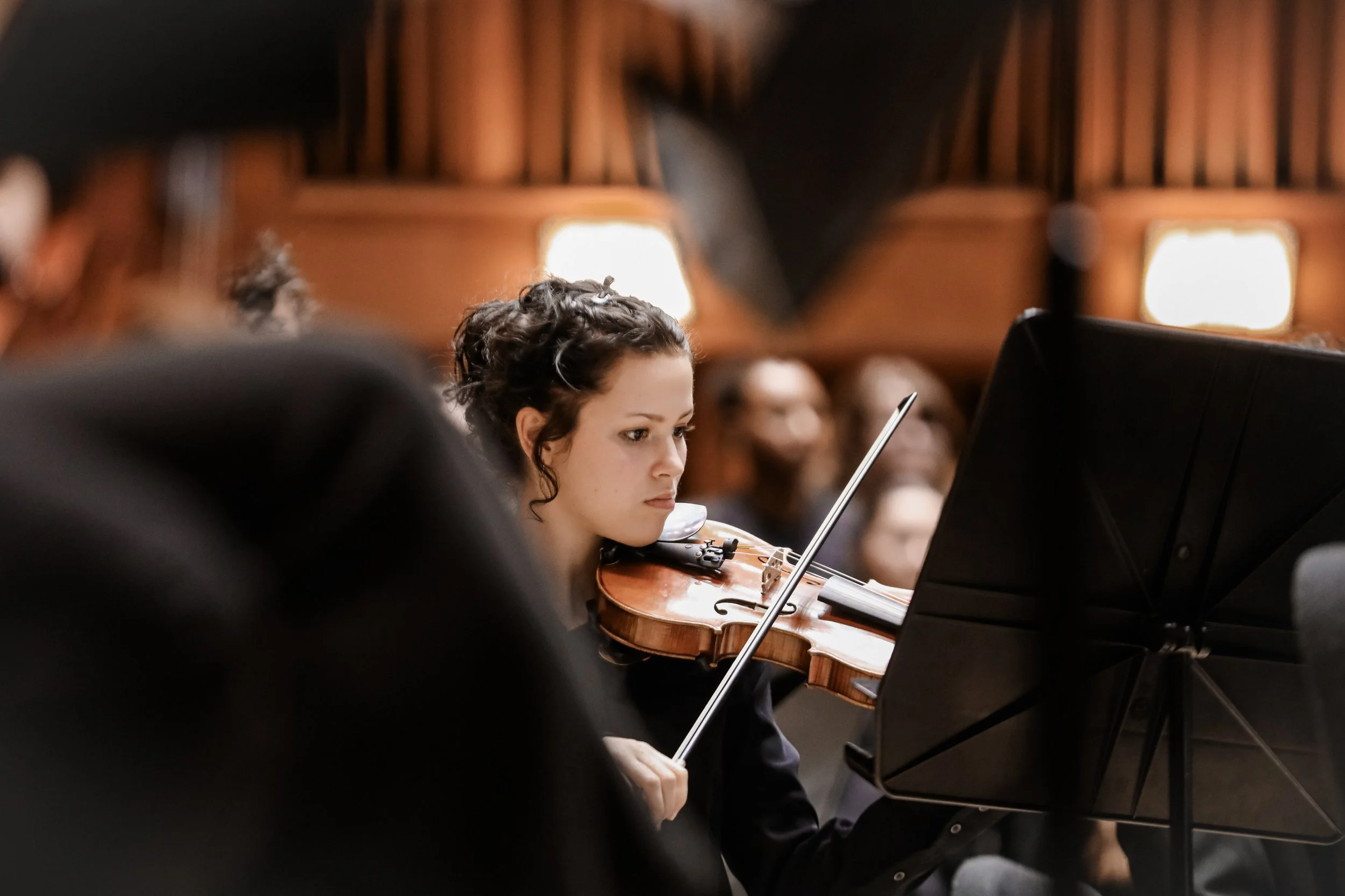 A young woman with curly hair playing the violin during an orchestra performance, with other orchestra members visible in the background.