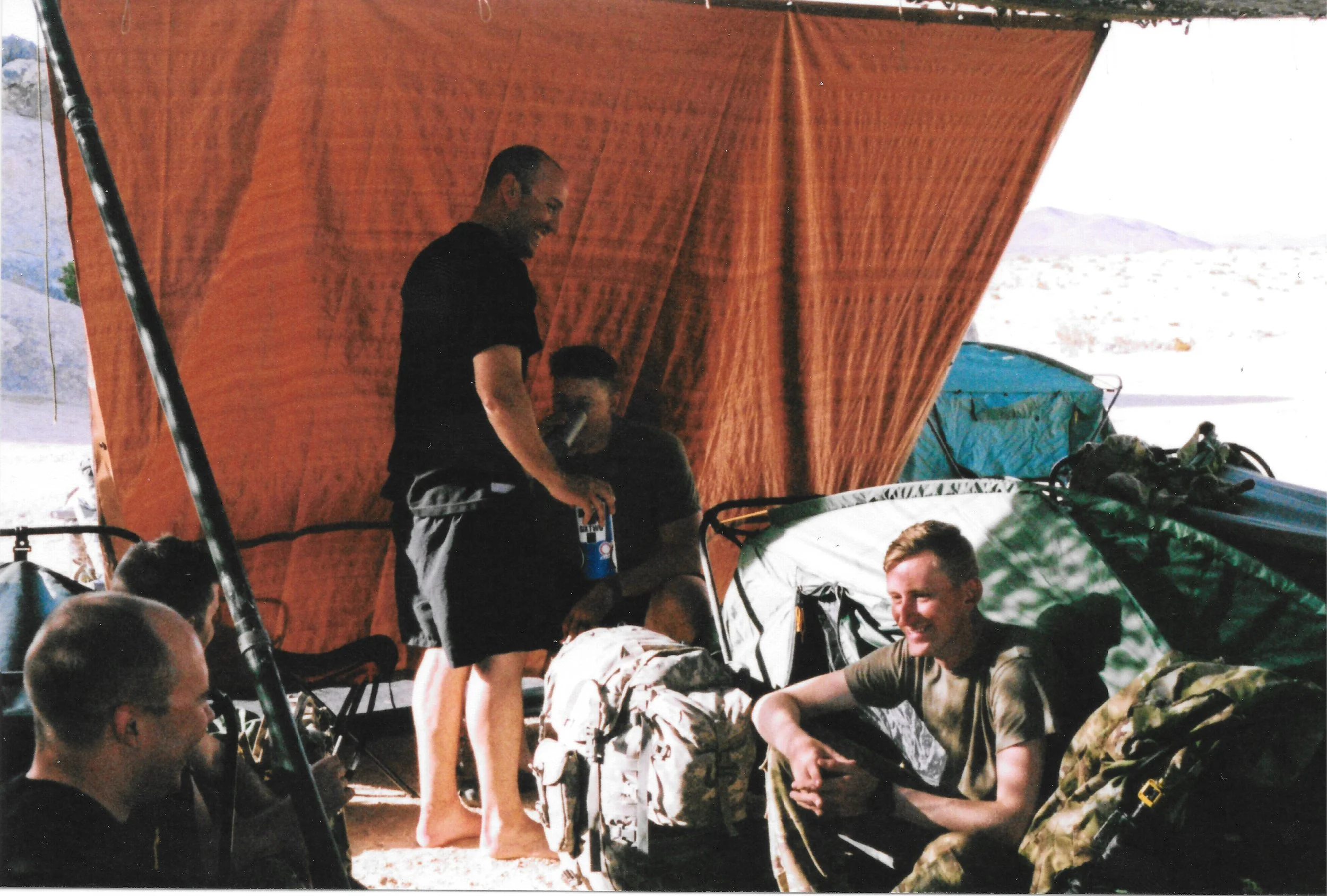 A group of people are gathered outdoors under a canopy, with some sitting and others standing. The scene appears to be relaxed and social, with a mountainous landscape in the background.