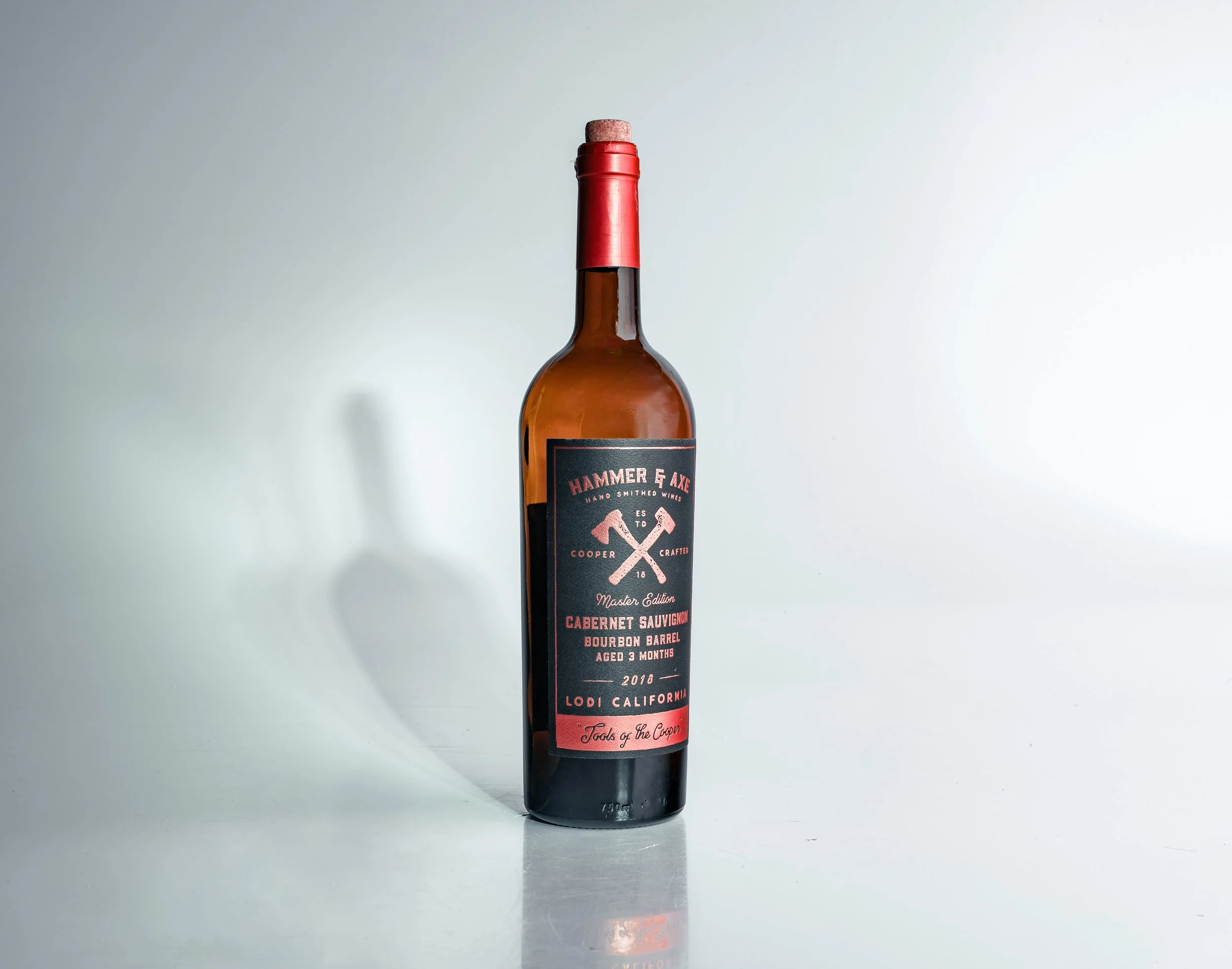 Bottle of Hammer & Axe Cabernet Sauvignon with a red cap and black label, placed on a reflective surface against a plain light background.