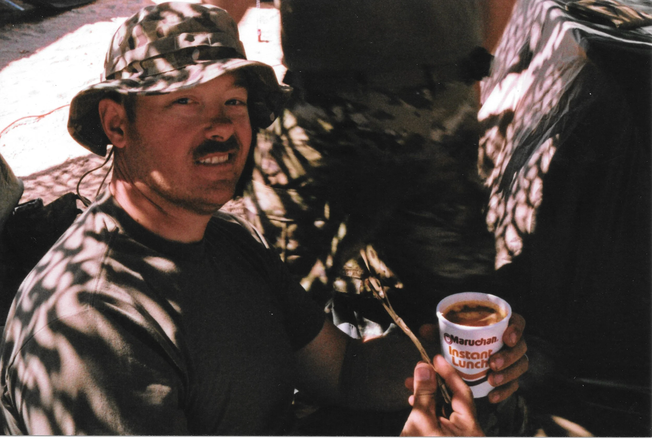 A man smiling and holding a cup of coffee, wearing a camouflage hat and jacket, seated at a table with a cup of coffee.