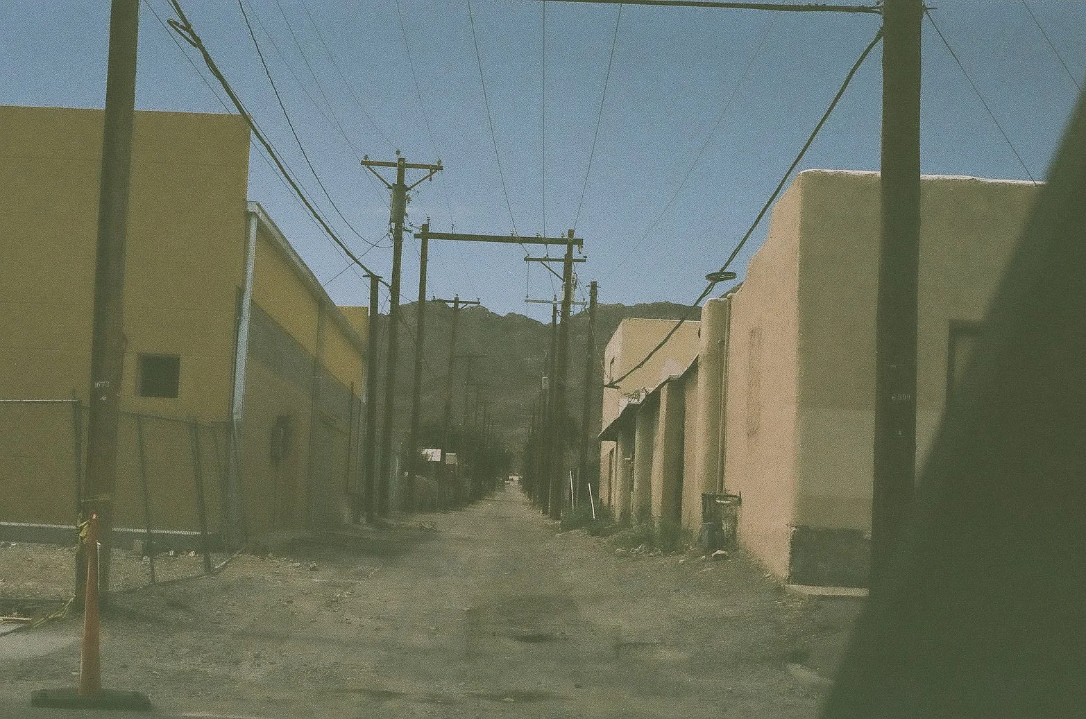 A narrow alleyway between buildings, with utility poles and wires overhead, in a desert area under clear blue sky.