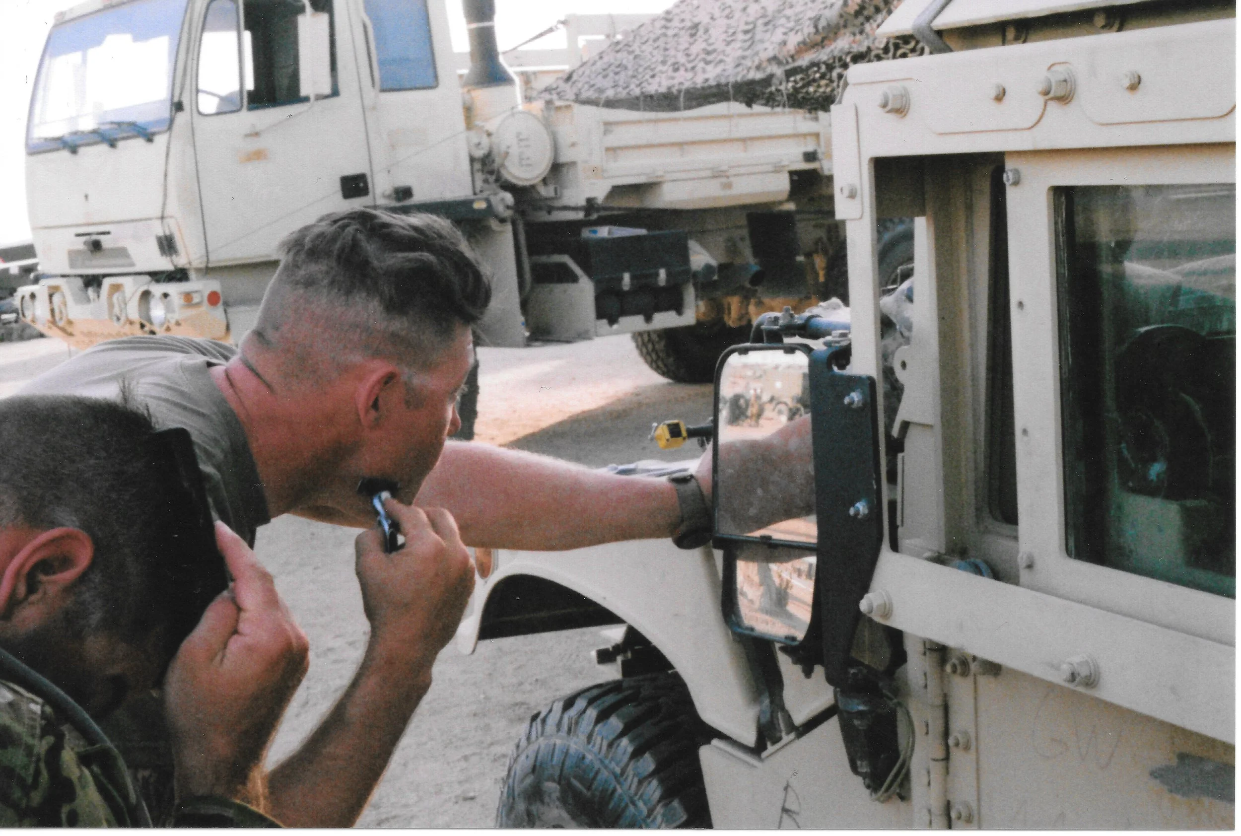 Two men working on a white vehicle, possibly an trucks, with one of them using a tool near the window, and a large piece of equipment or cargo in the background.
