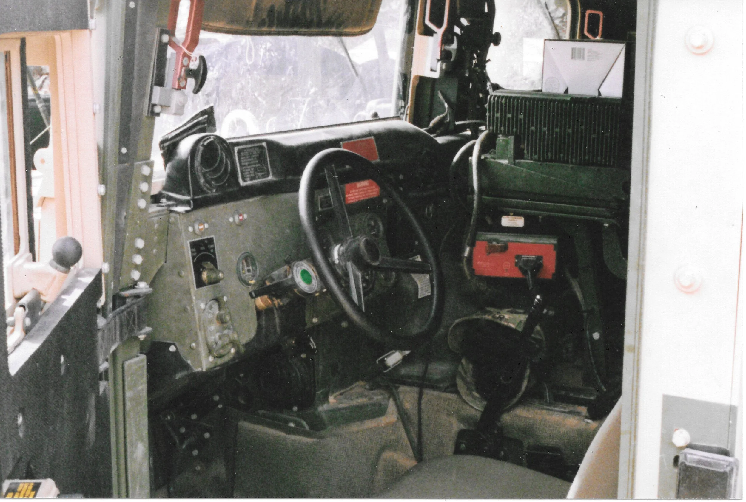 The image shows the interior of a cockpit or control station, possibly in an aircraft or simulator. It contains various control panels, gauges, switches, and a steering wheel or control stick. The view outside the window shows an indistinct outdoor s