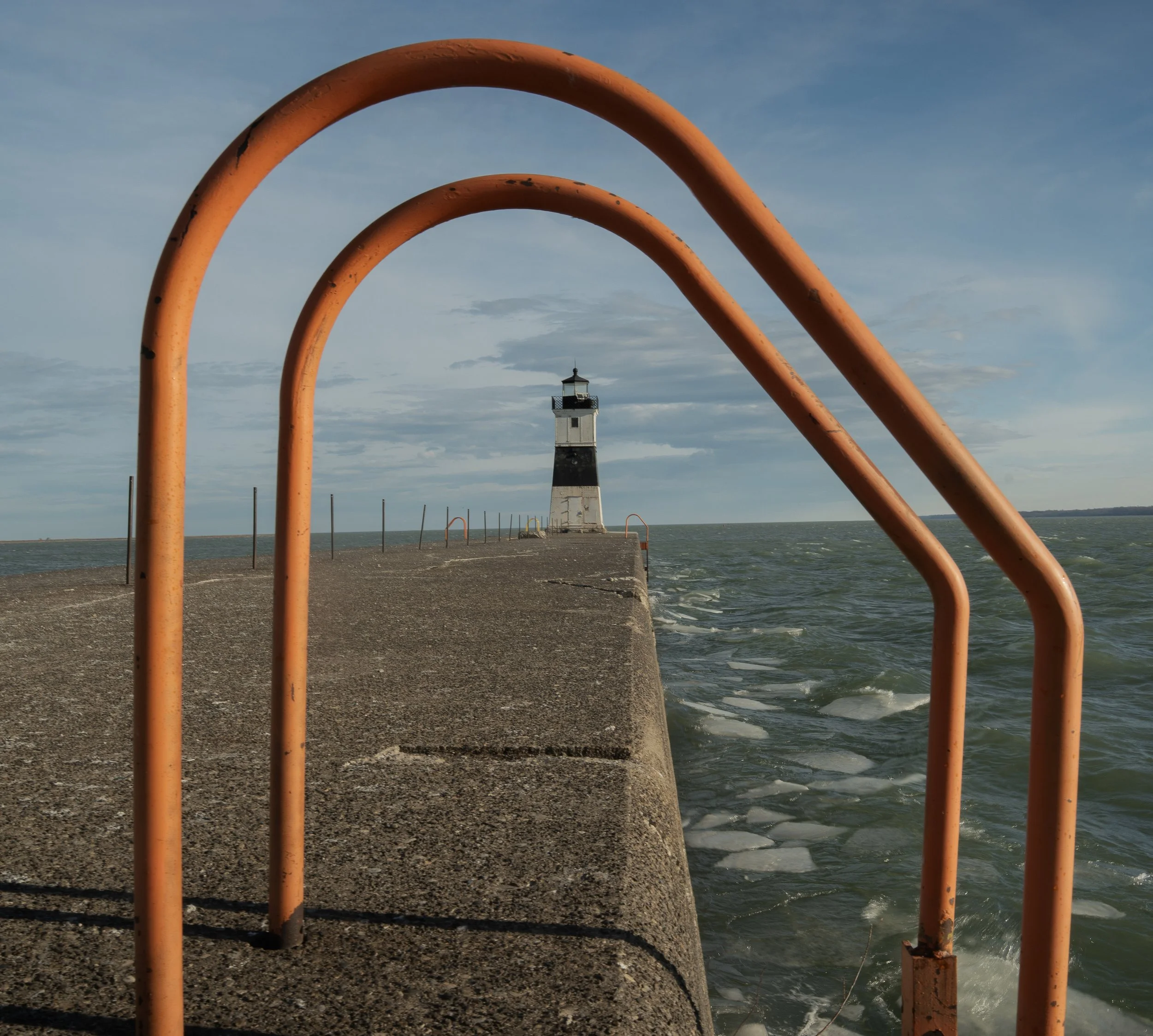 Orange railing in the foreground with a lighthouse in the background on a beach with water and sky.
