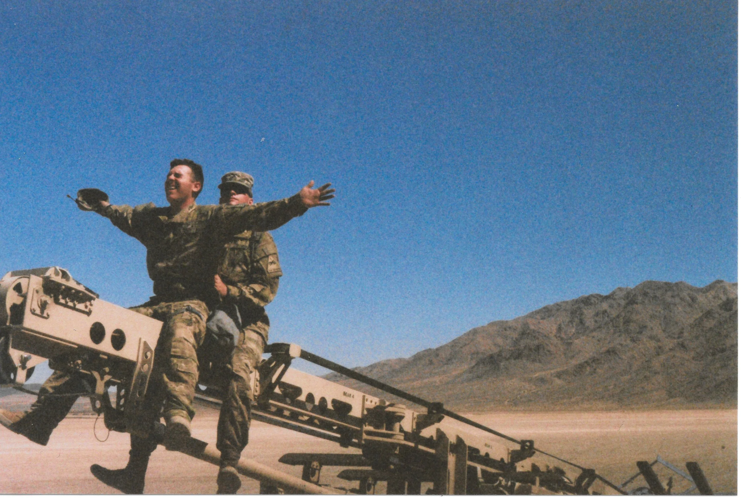 Two soldiers in camouflage uniforms are boarding a military vehicle with a ramp, in a desert landscape with mountains and a clear blue sky.