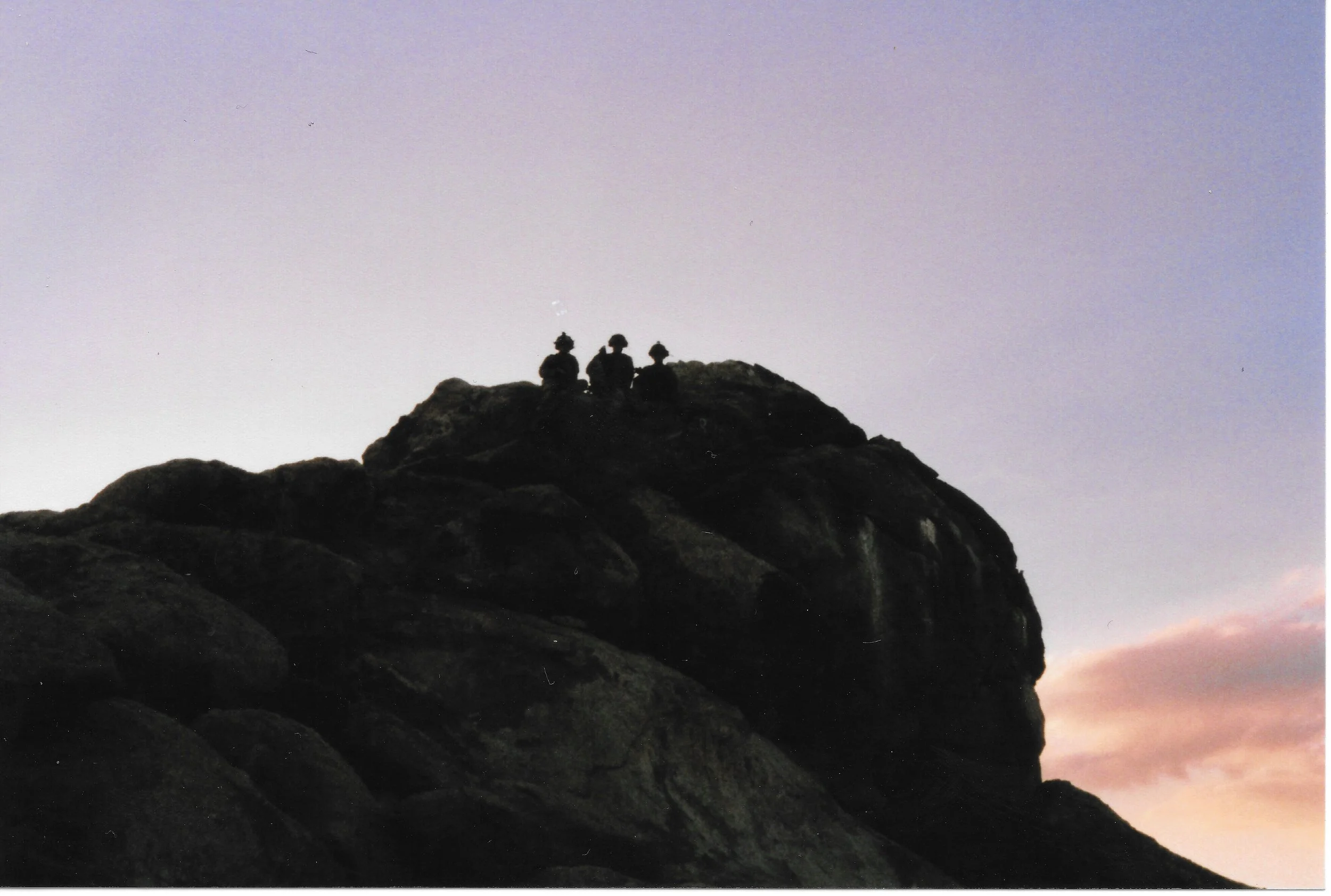 Silhouettes of three soldiers sitting on a rocky hilltop during sunset.