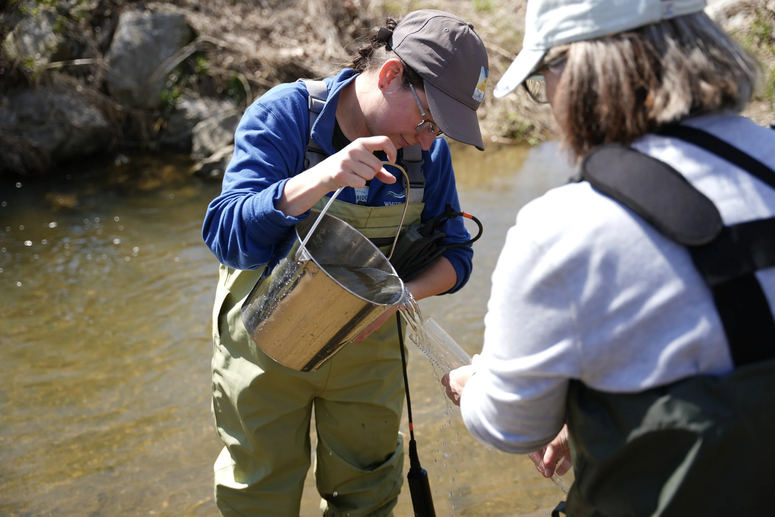 Two people in outdoor gear examining water flow and holding a measuring device in a creek.