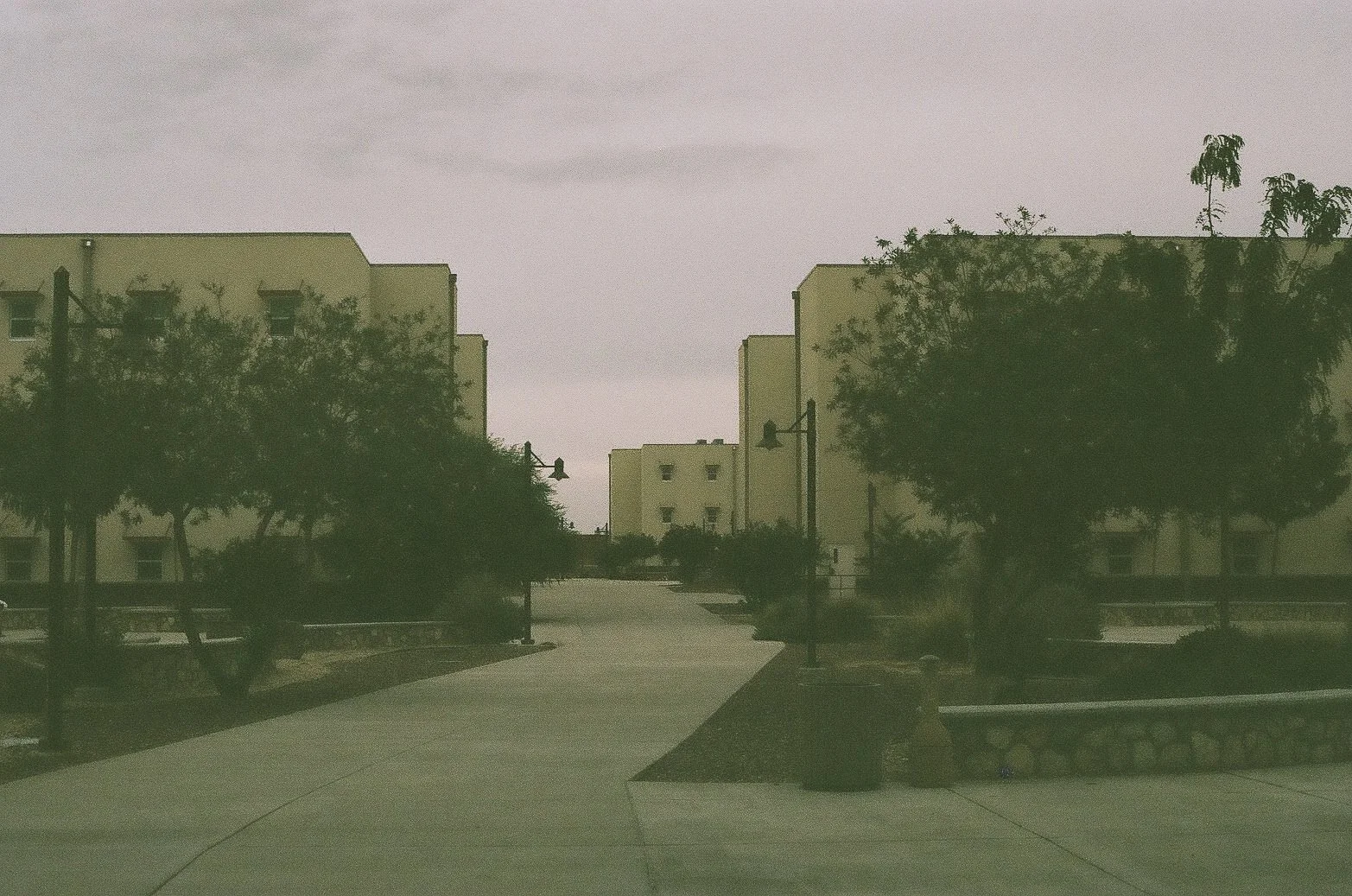 A residential area with low-rise apartment buildings, trees, street lamps, and a paved walkway in the foreground.