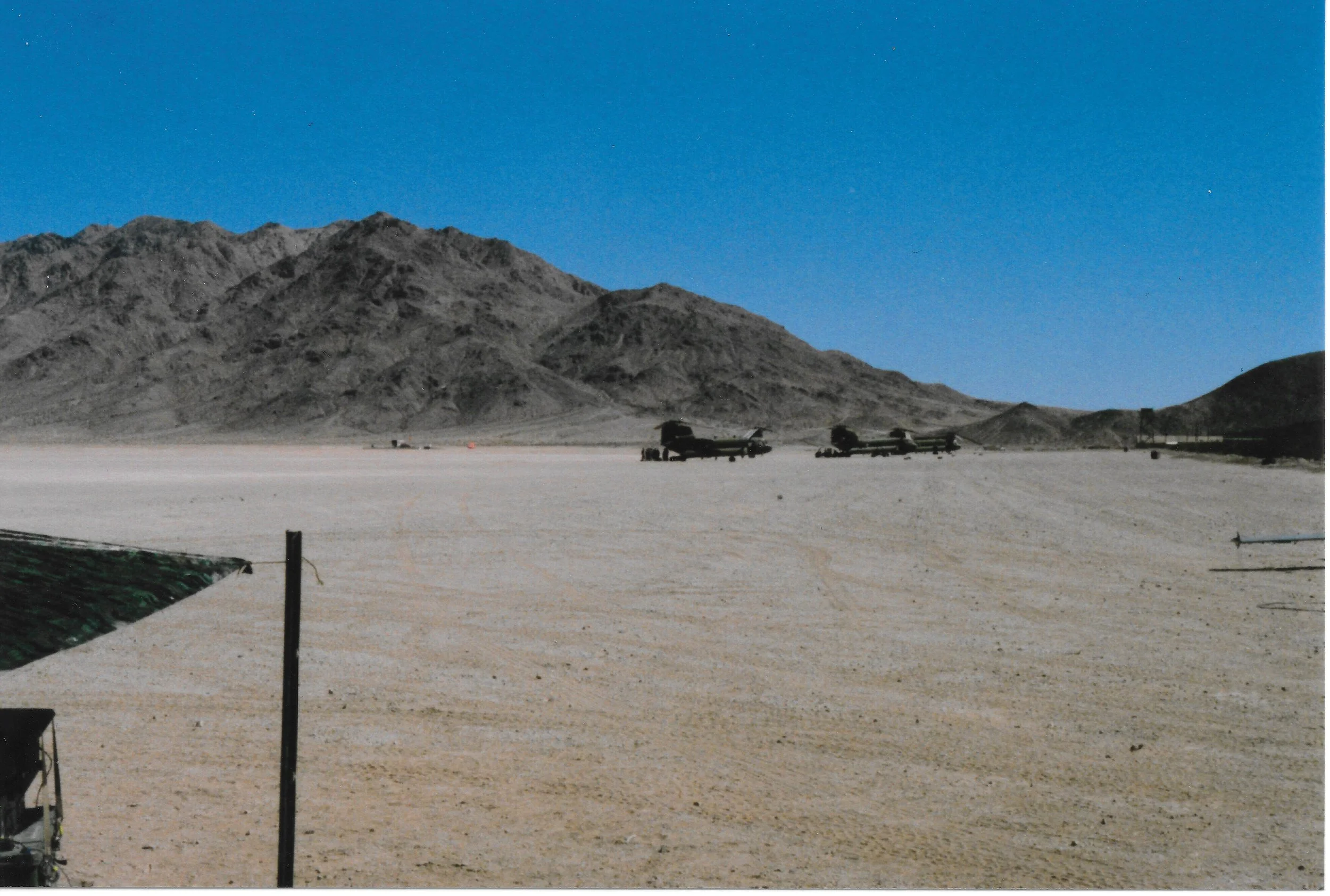 Desert landscape with mountains in the distance, a clear blue sky, and several military vehicles parked on the sandy ground in the foreground.