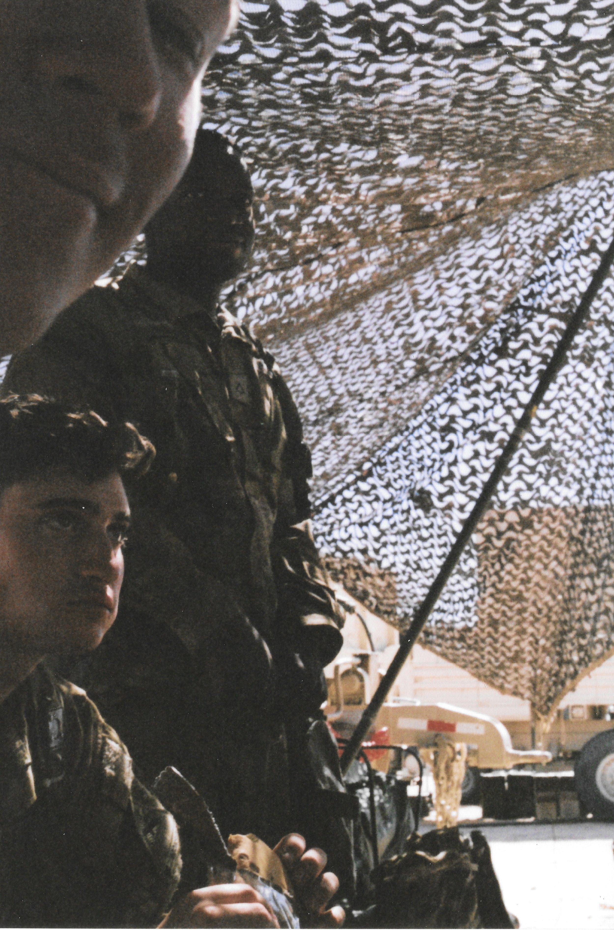 Three armed soldiers in camouflage uniforms standing outdoors in front of a camouflage net, with a military vehicle and some equipment visible in the background.