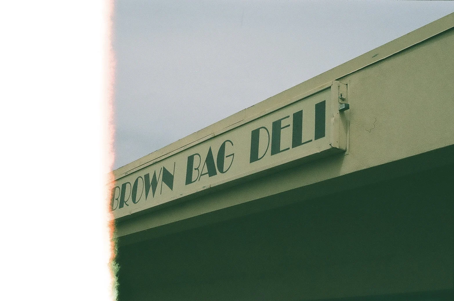 Close-up of a sign that reads "BROWN BAG DELI" on a building exterior, with a light blue sky in the background and a partial green awning below.