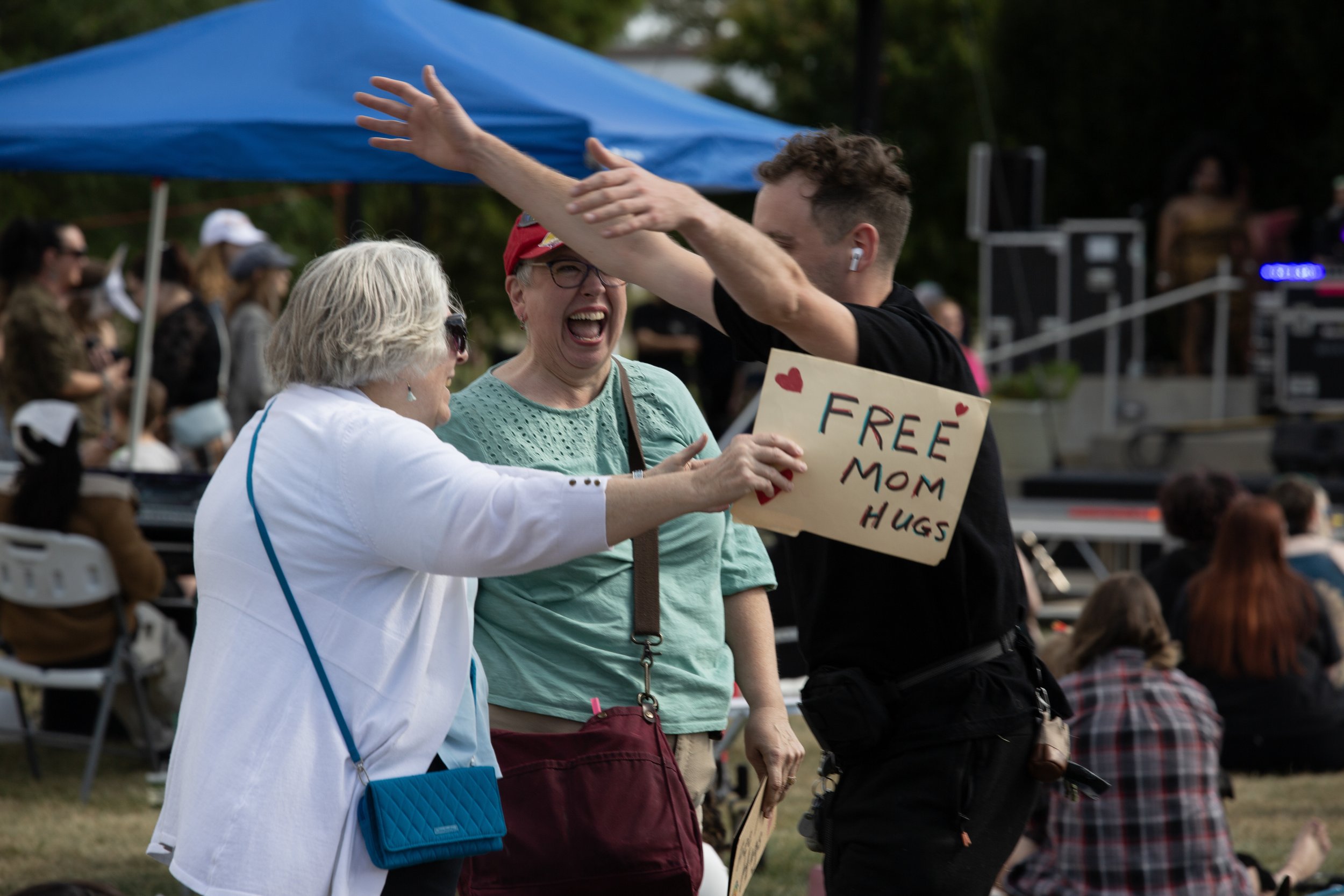 Three women in a public outdoor gathering, smiling and hugging. The woman on the right is holding a sign that reads 'FREE MOM HUGS' decorated with hearts. The background shows organized seating and a blue canopy, with more people present.