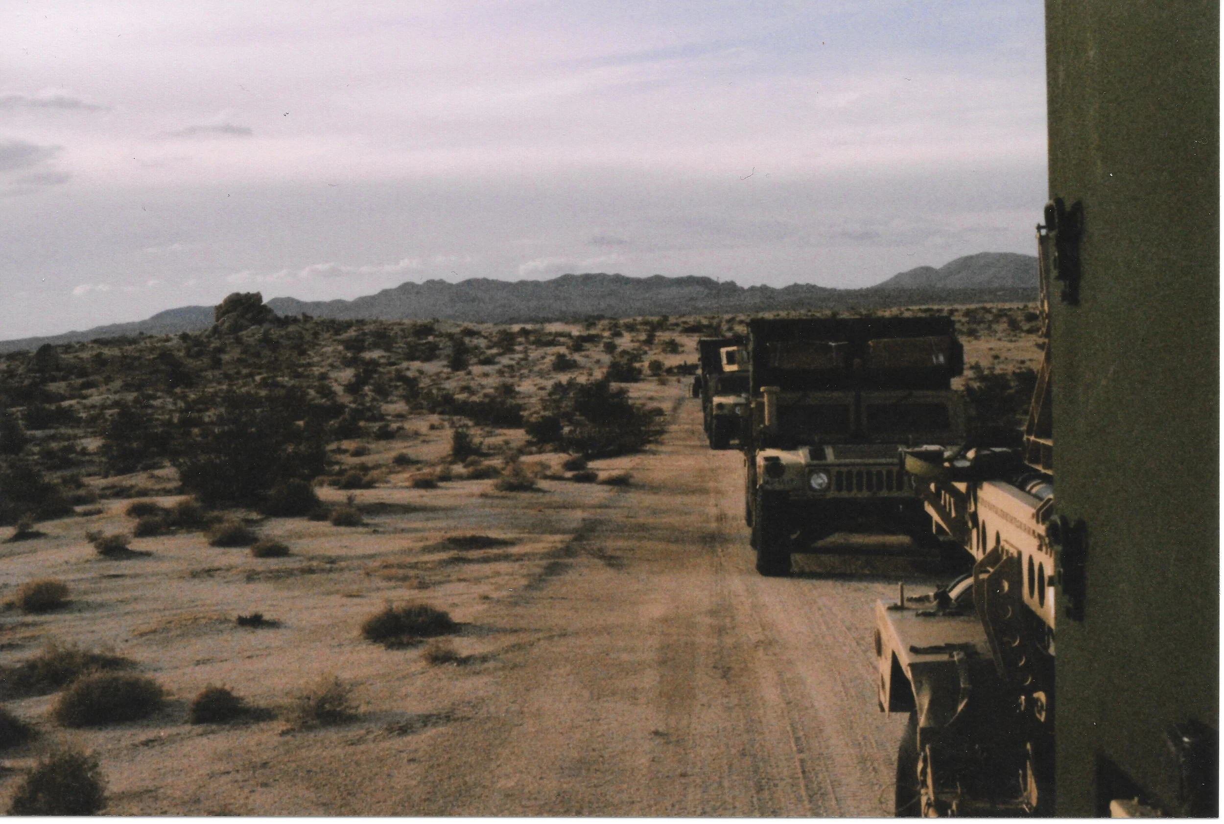 Desert landscape with mountains in the distance, two trucks on a dirt road, and a large structure or wall on the right side of the image.