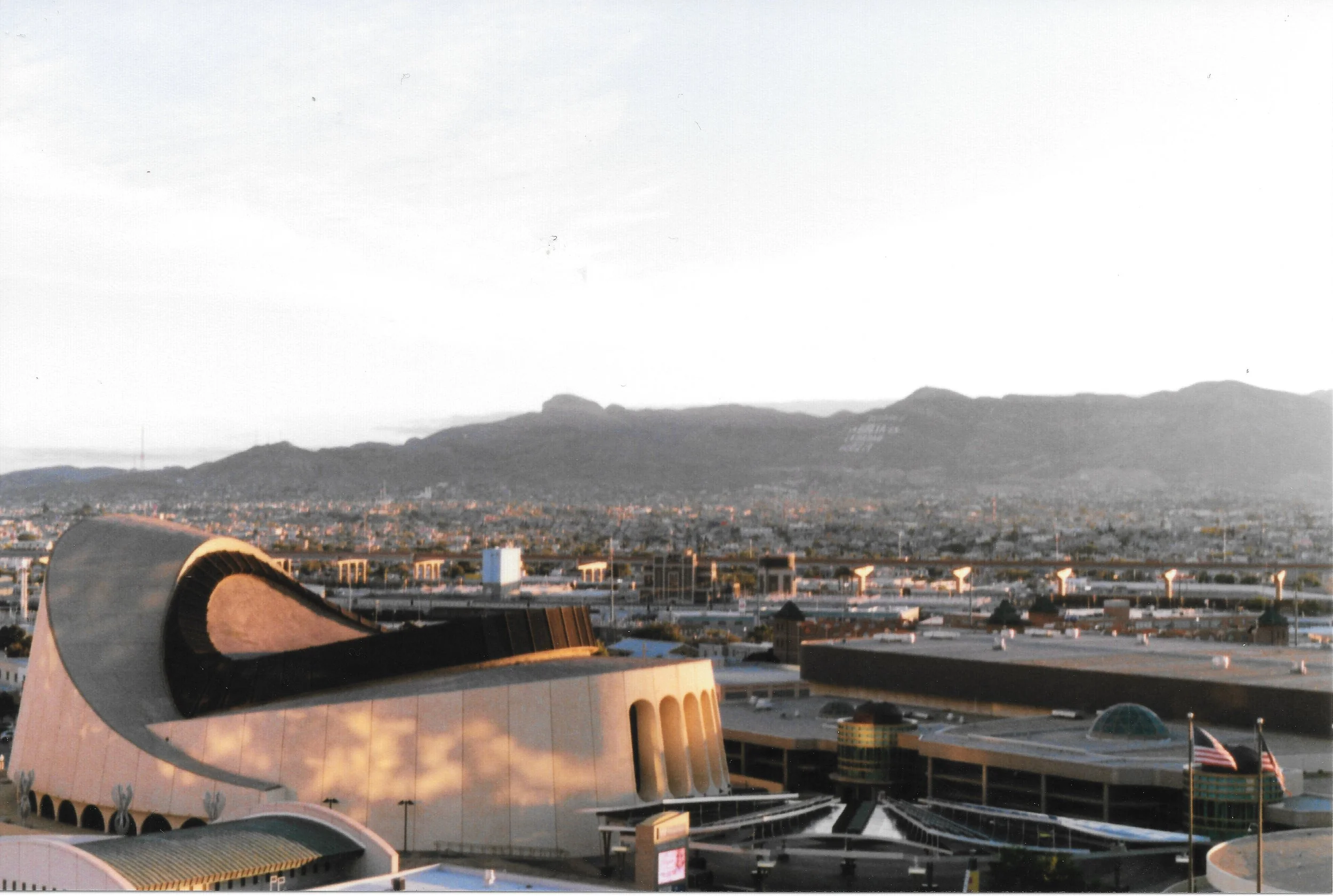 View of a cityscape with a modern building in the foreground, mountains in the background, and a clear sky.