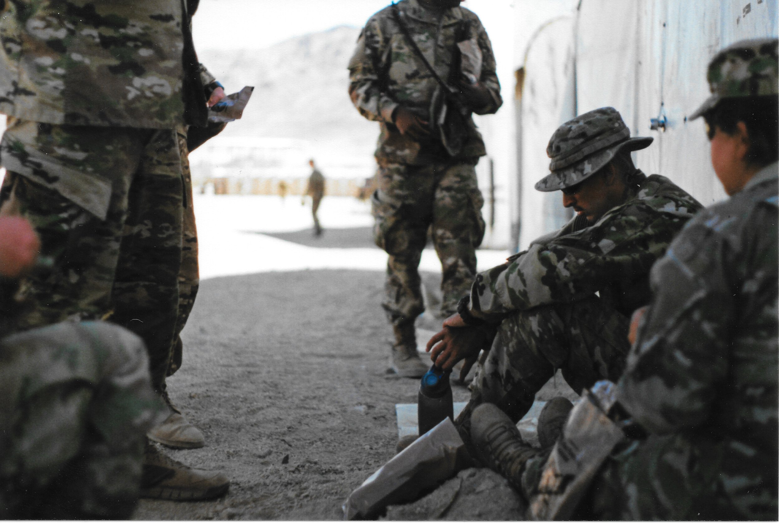 Military personnel in camouflage uniforms sitting on the ground and standing against a wall, with some holding water bottles, in a cold, snowy environment.