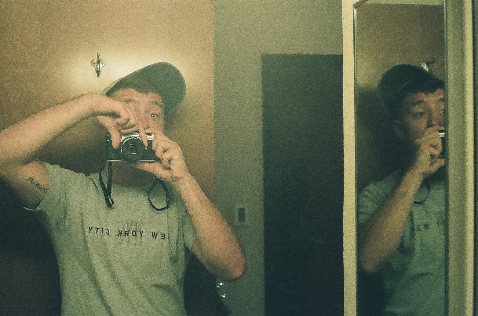 A young man wearing a cap and a gray t-shirt taking a selfie in front of a mirror, holding a camera up to his face. His reflection is visible in the mirror, which is mounted on a door or wall.