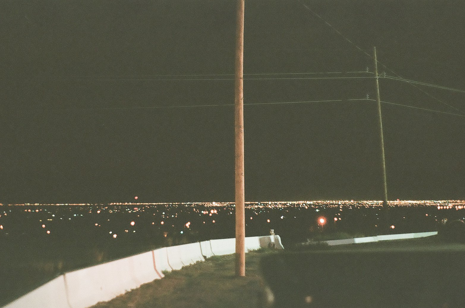 Nighttime cityscape view with illuminated city lights, a utility pole in the foreground, and a winding road with guardrails.