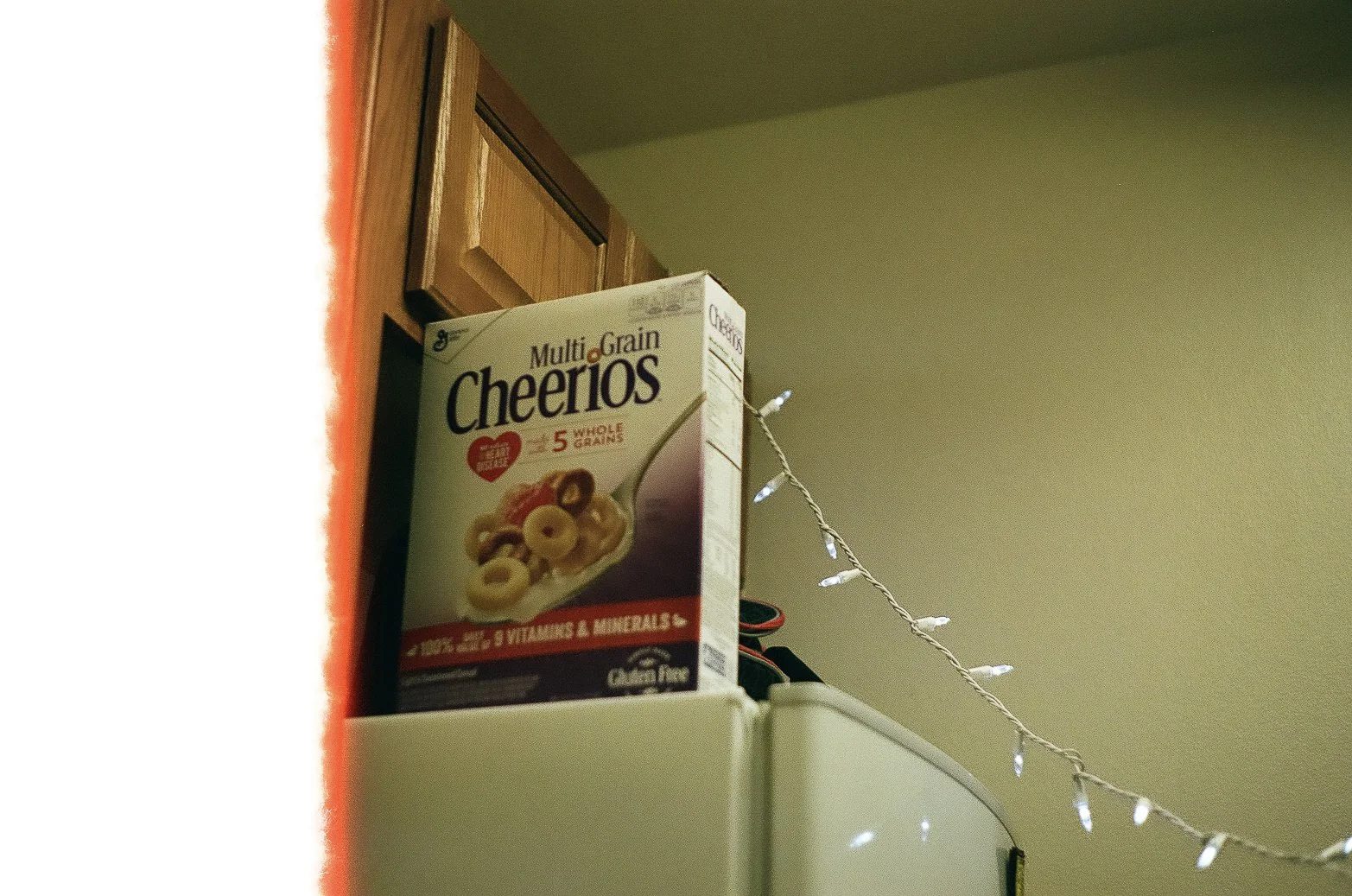 A box of Multi Grain Cheerios cereal on top of a white appliance with a string of white fairy lights hanging nearby and a wooden cabinet in the background.