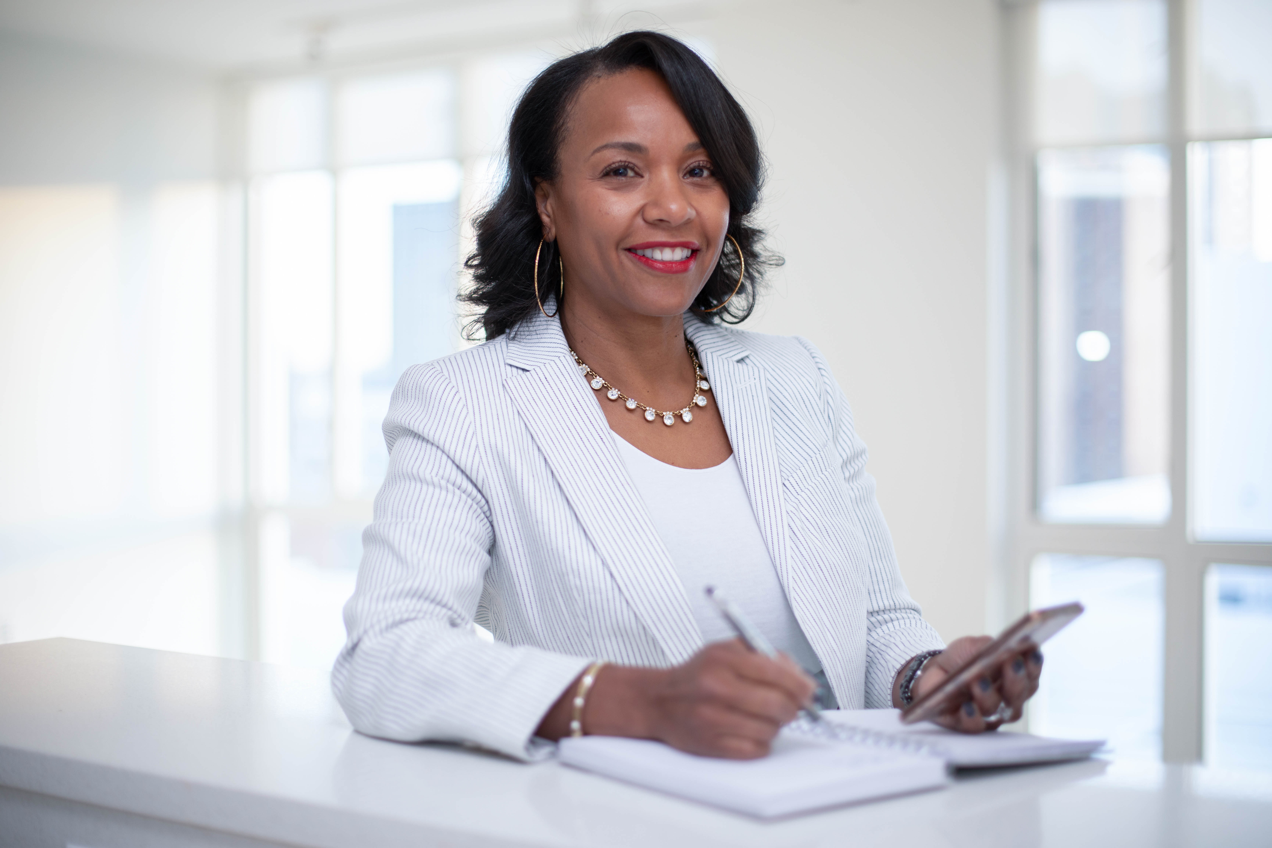 A woman with dark hair, wearing a white blazer and jewelry, sitting at a white table, smiling, holding a smartphone with windows in the background.