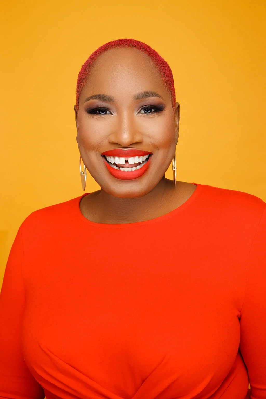 A woman with short red hair, bright red lipstick, and hoop earrings, wearing a bright orange top, smiling against a yellow background.