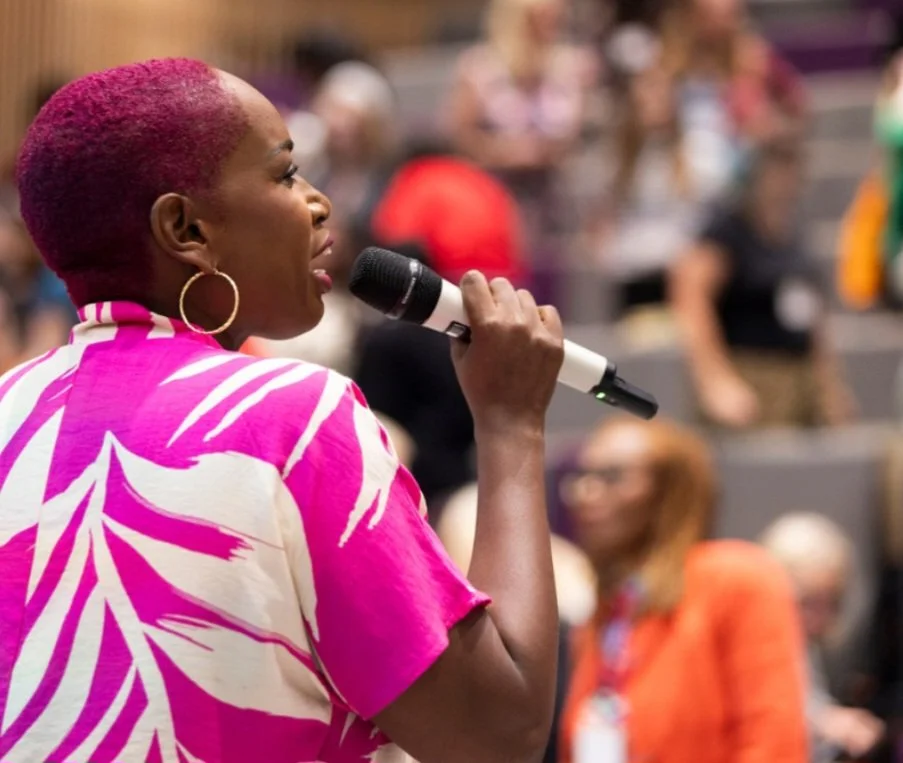 A woman with short pink hair and hoop earrings speaking into a microphone at an event with a blurred audience in the background.