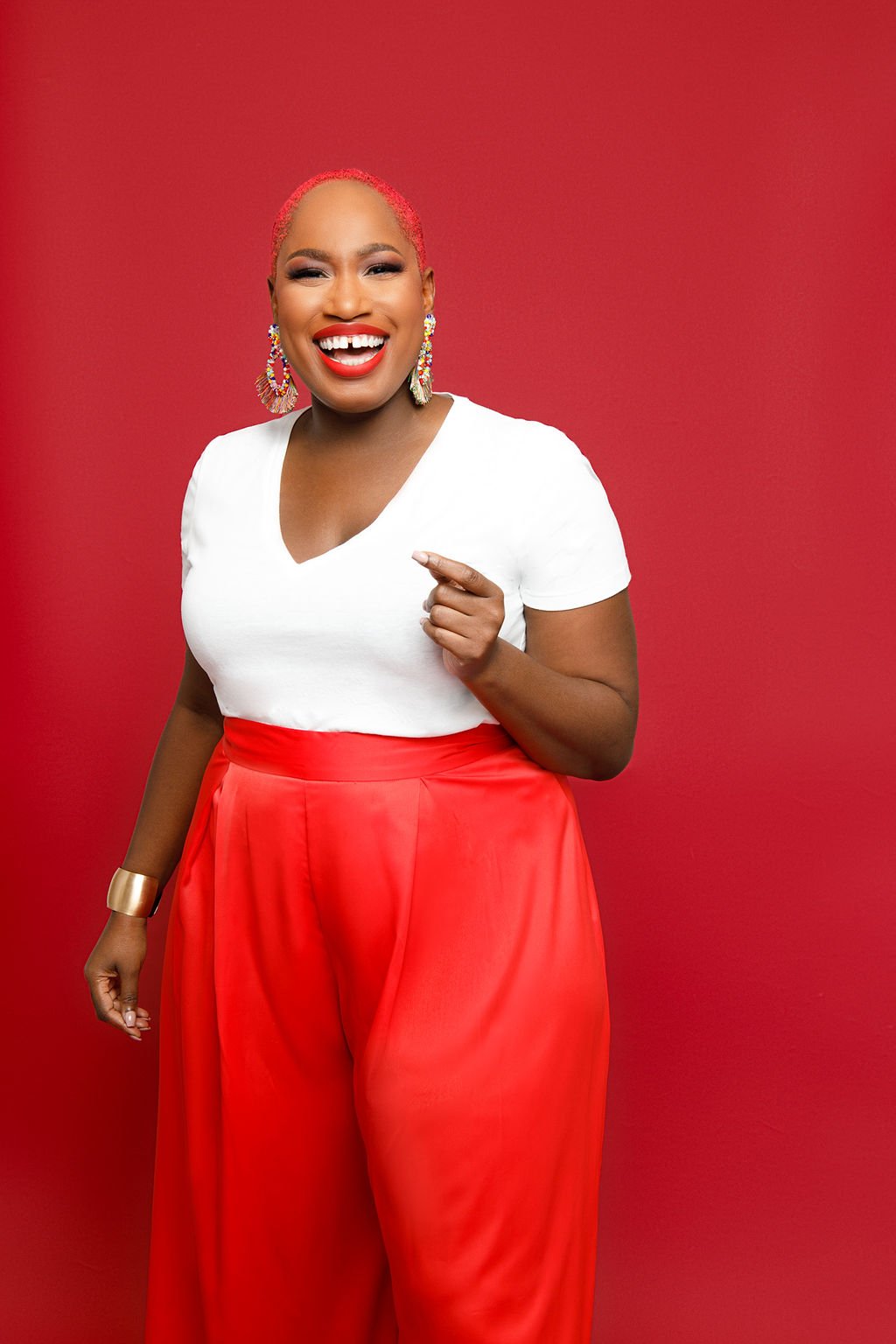 A woman with short pink hair, wearing a white top and red wide-leg pants, smiling and pointing, standing against a red background.