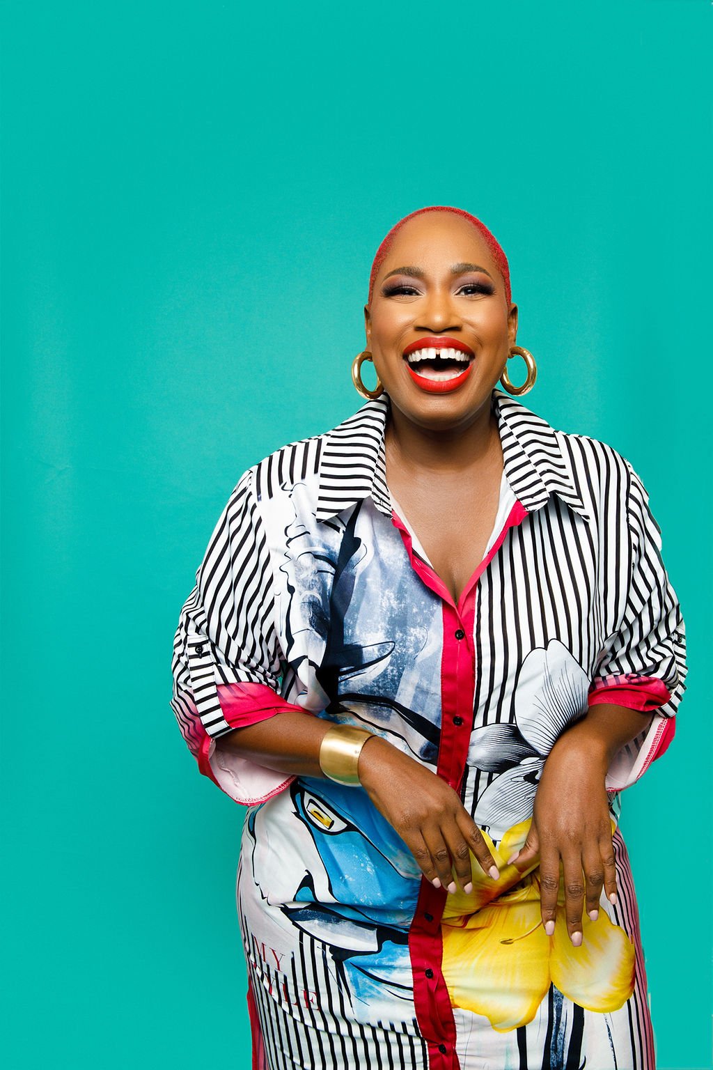 A woman with short red hair, big hoop earrings, bright red lipstick, and a striped, colorful dress smiling against a teal background.