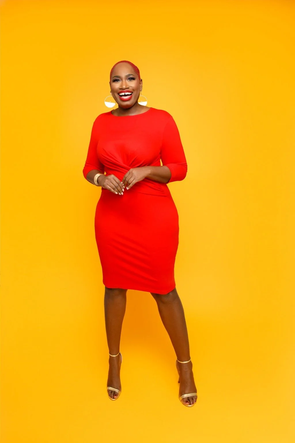 A woman with short red hair, wearing a red dress, smiling, standing against a yellow background.