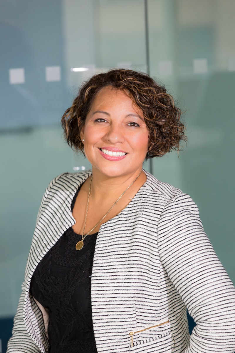 A smiling woman with curly hair wearing a black top and a striped blazer, standing indoors in front of glass panels.