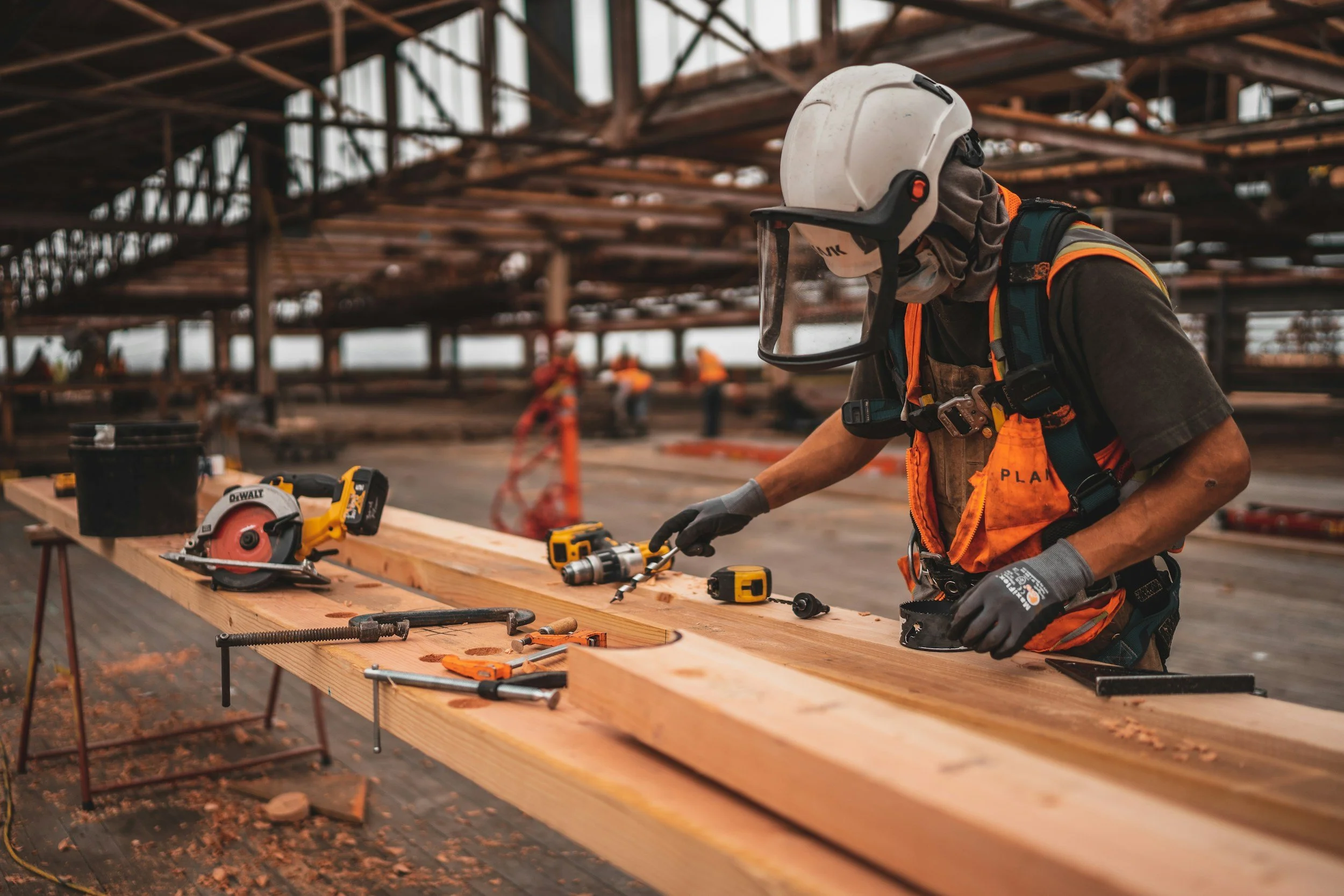 Operaio indossa casco, guanti e giacca ad alta visibilità mentre lavora con strumenti su un lungo pezzo di legno in un cantiere edile.
