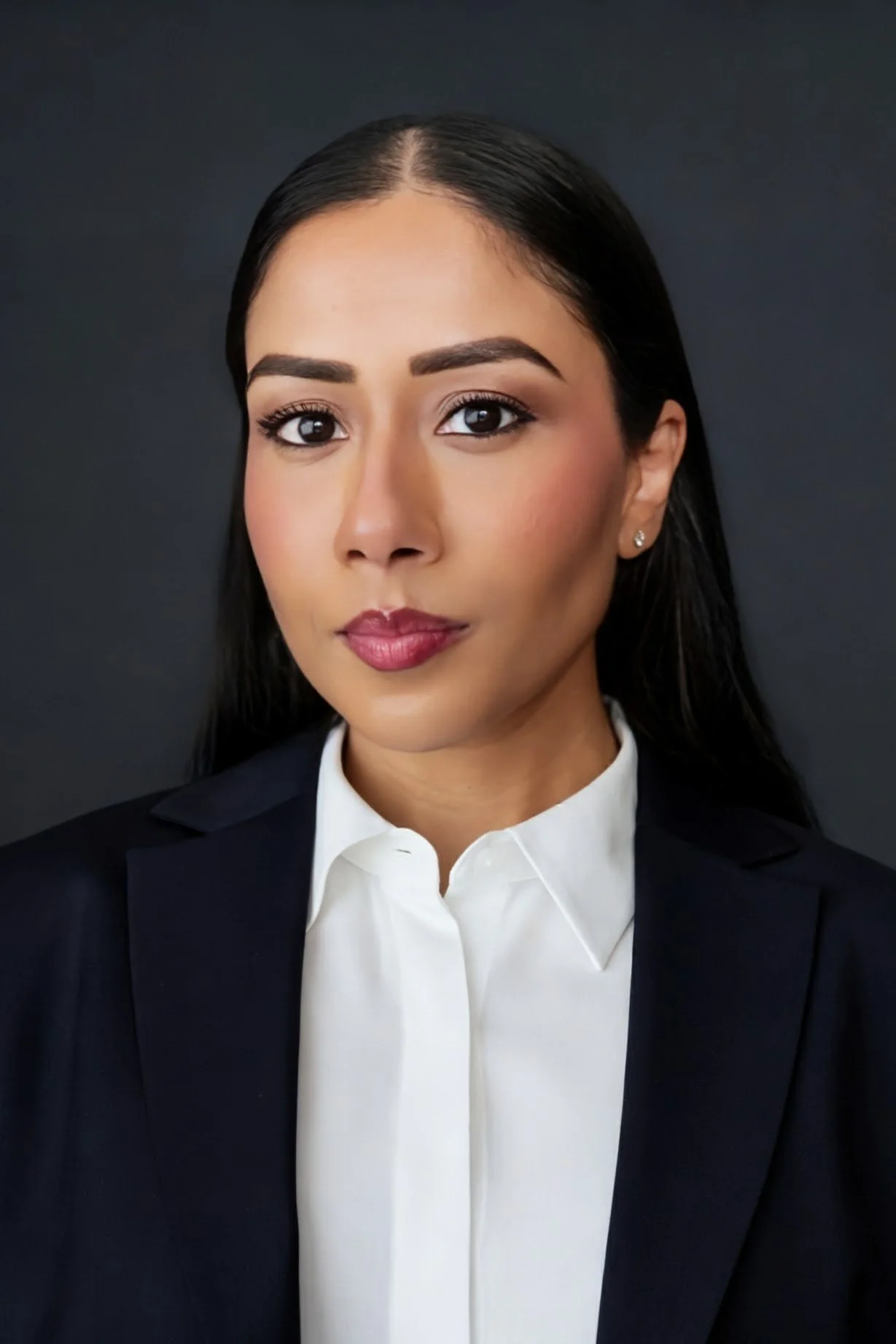 A professional woman with dark hair, makeup, and earrings, wearing a black blazer and white shirt, against a dark background.