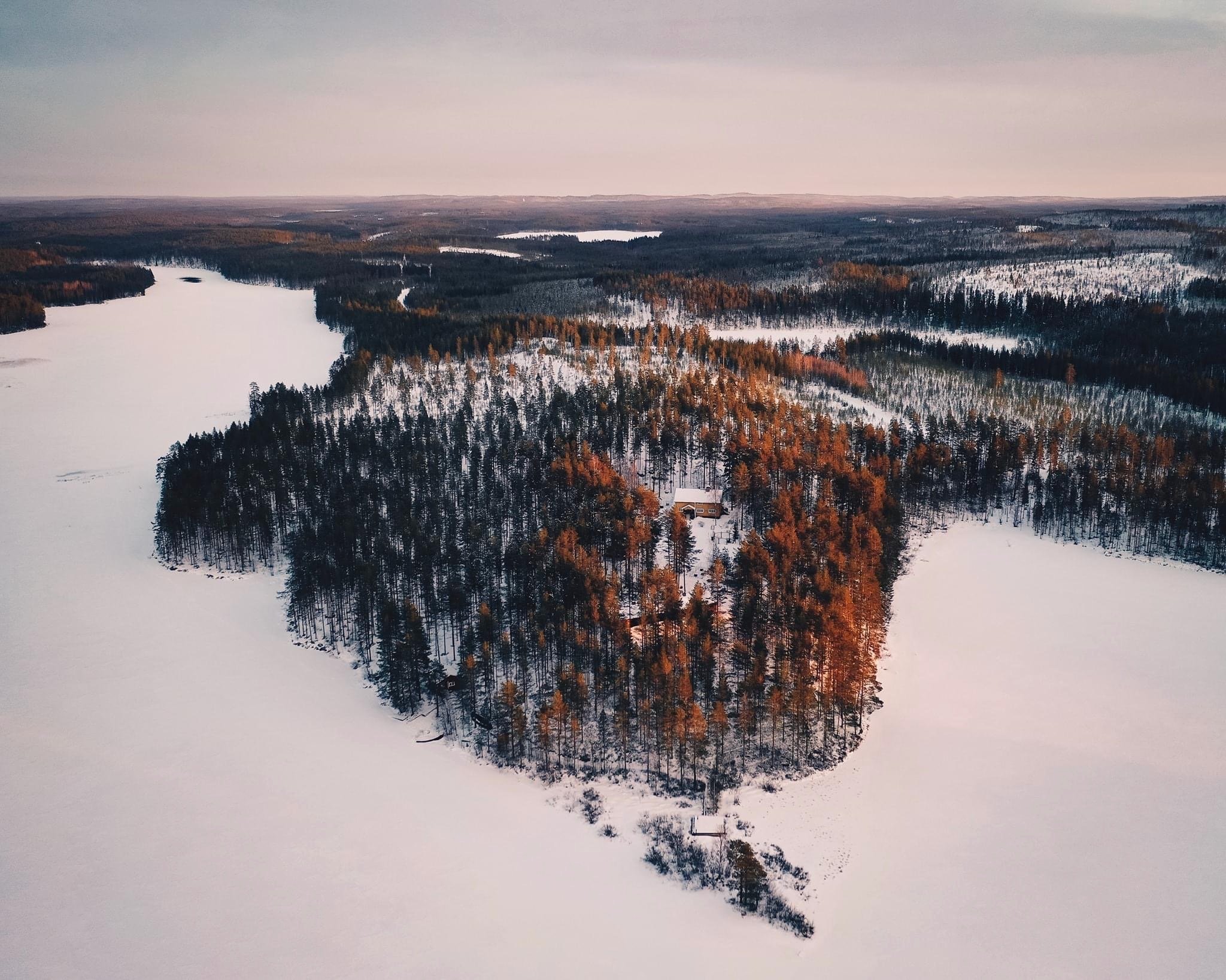 Aerial view of the snowy landscape of Kokkojärvi with a dense forested island and a house, surrounded by frozen water, under a cloudy sky.
