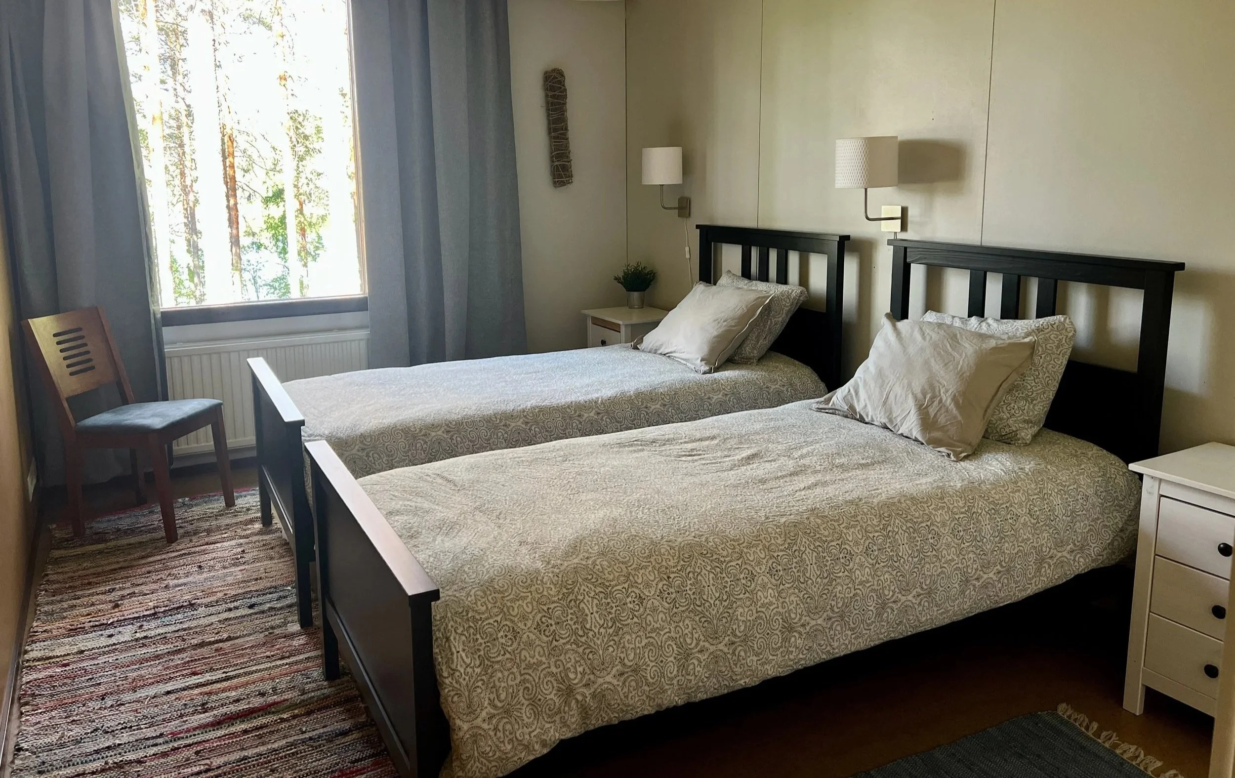 Two twin beds with black wooden headboards, beige patterned bedding, and pillows, in a hotel room with a window, a nightstand with a potted plant, wall-mounted lamps, a wooden chair, curtains, and a rug on the floor.