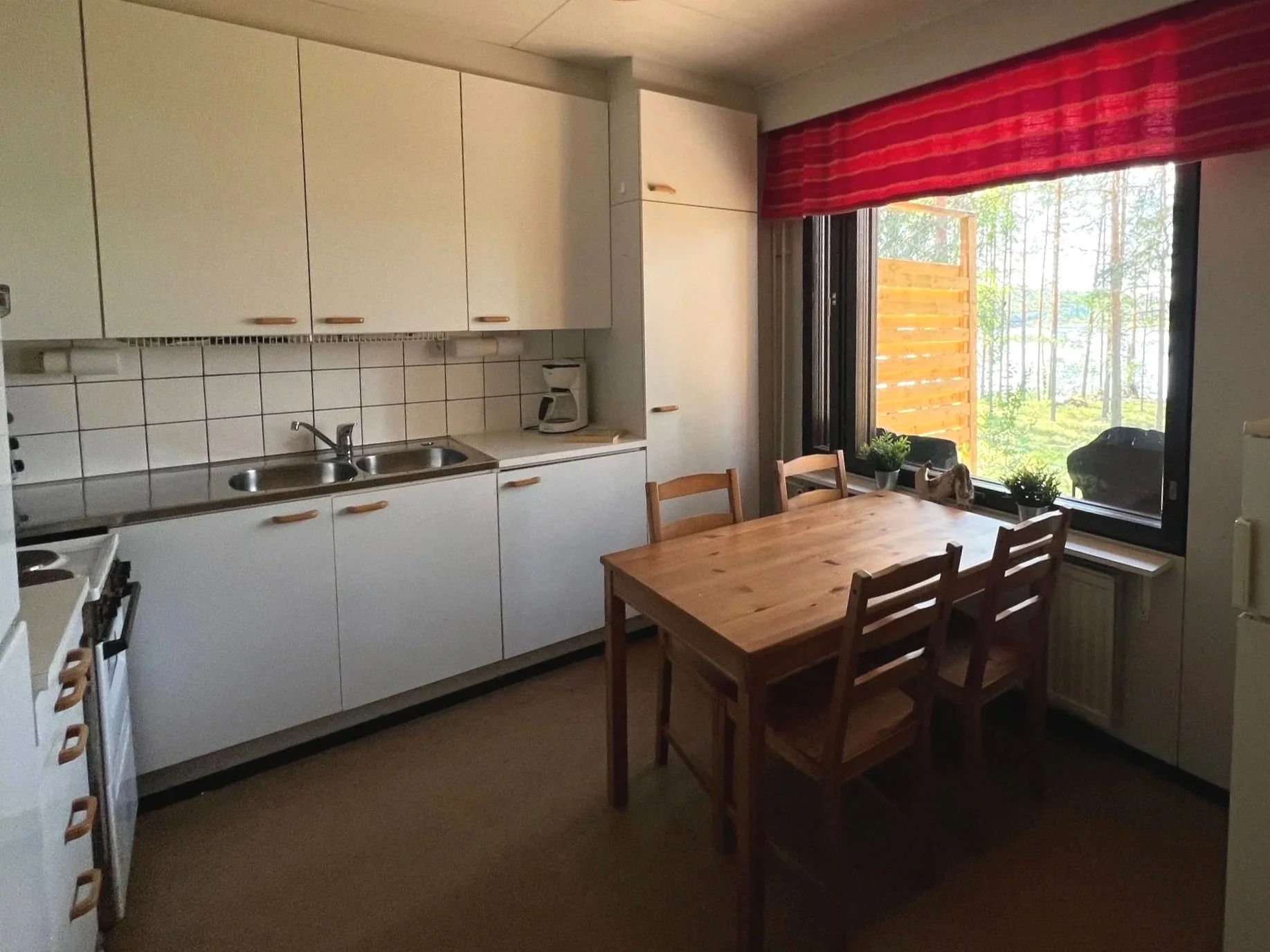 A simple kitchen with white cabinets and a wooden dining table near a large window overlooking a forested area, with potted plants on the windowsill.